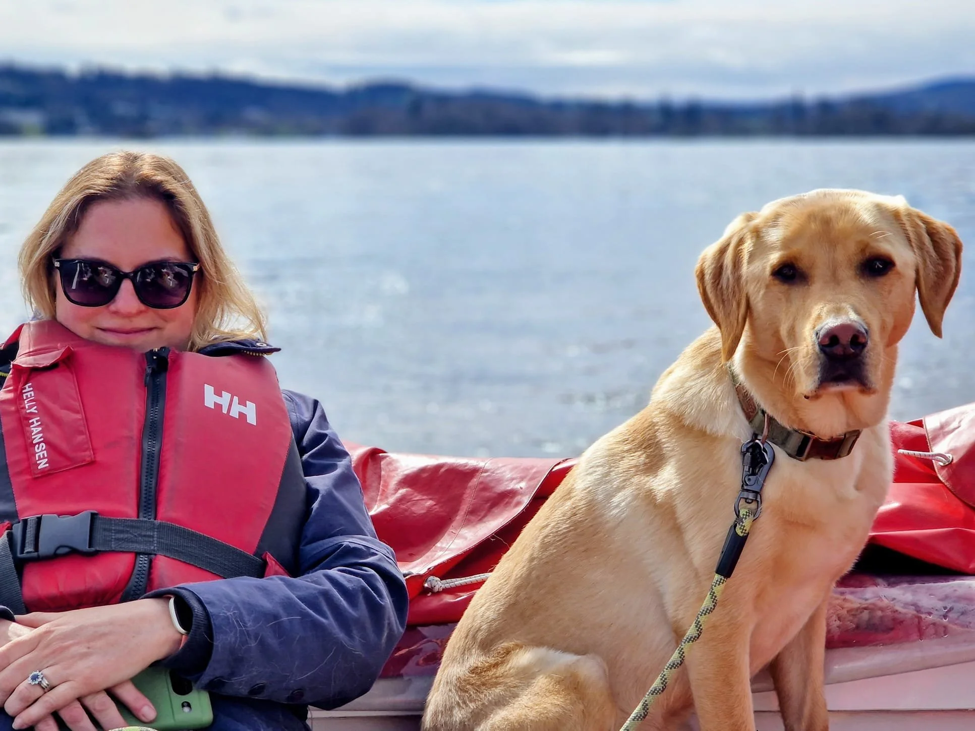 A woman wearing sunglasses and a red life jacket sitting on a boat with a yellow Labrador retriever dog in front of her, near a body of water with distant shoreline and mountains in the background.