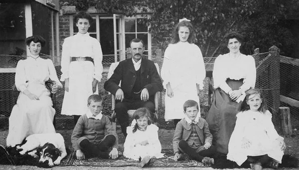 A black-and-white photo of seven children and one man, possibly a family, seated and standing outdoors in front of a house with a fence and trees.