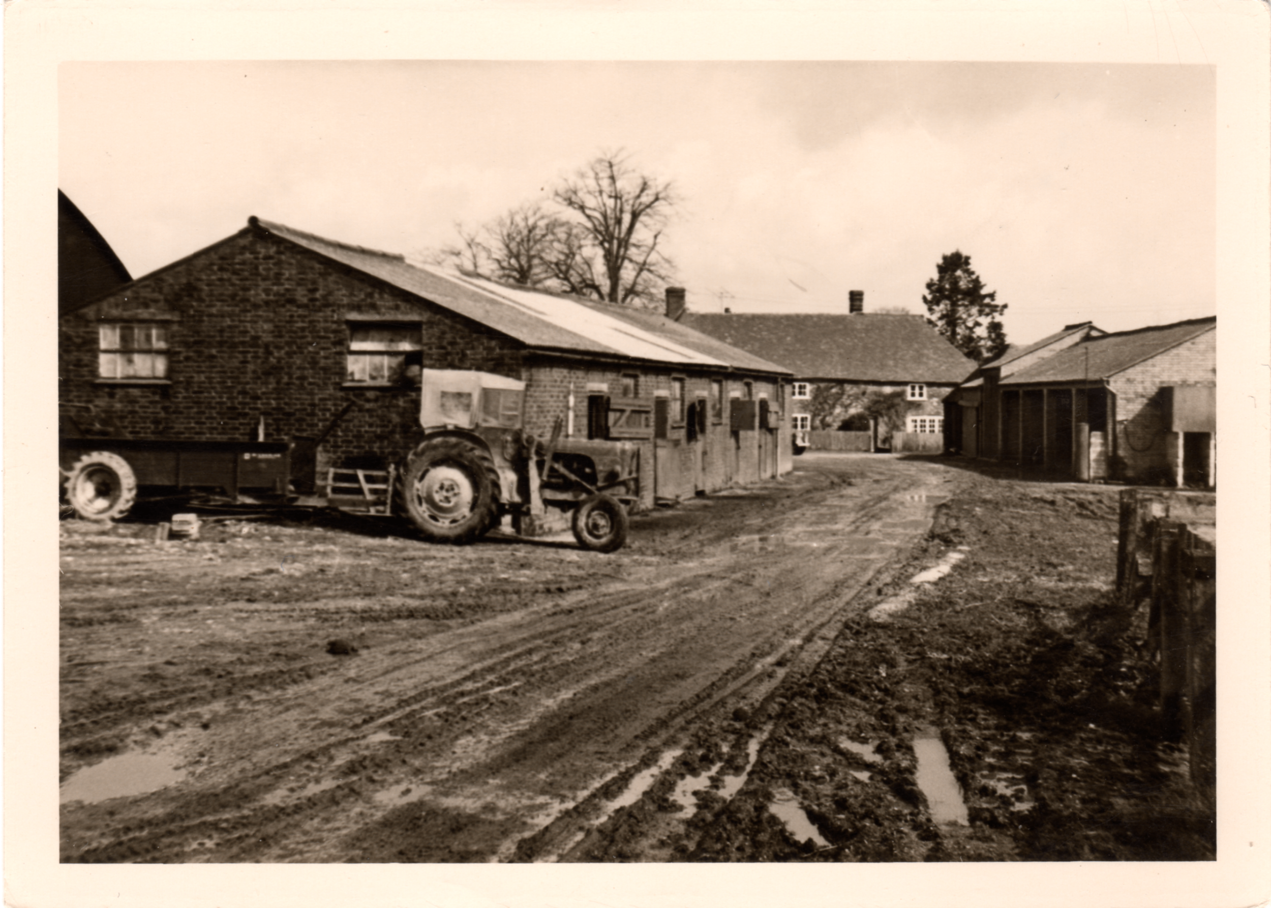 A black and white photograph of a rural scene featuring a tractor parked on a dirt road near brick houses with tiled roofs and a large tree with no leaves in the background.