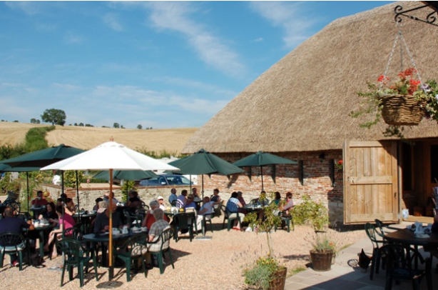 People dining outdoors with umbrellas in front of a rustic building with a thatched roof at a farm or countryside venue.