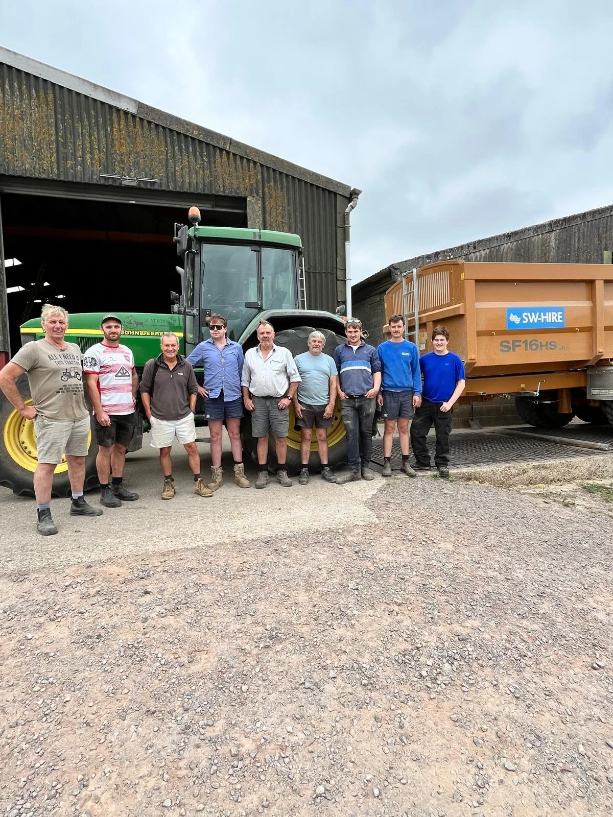 A group of ten men standing in front of a large green tractor and a brown trailer, outside a barn or garage on a gravel surface.