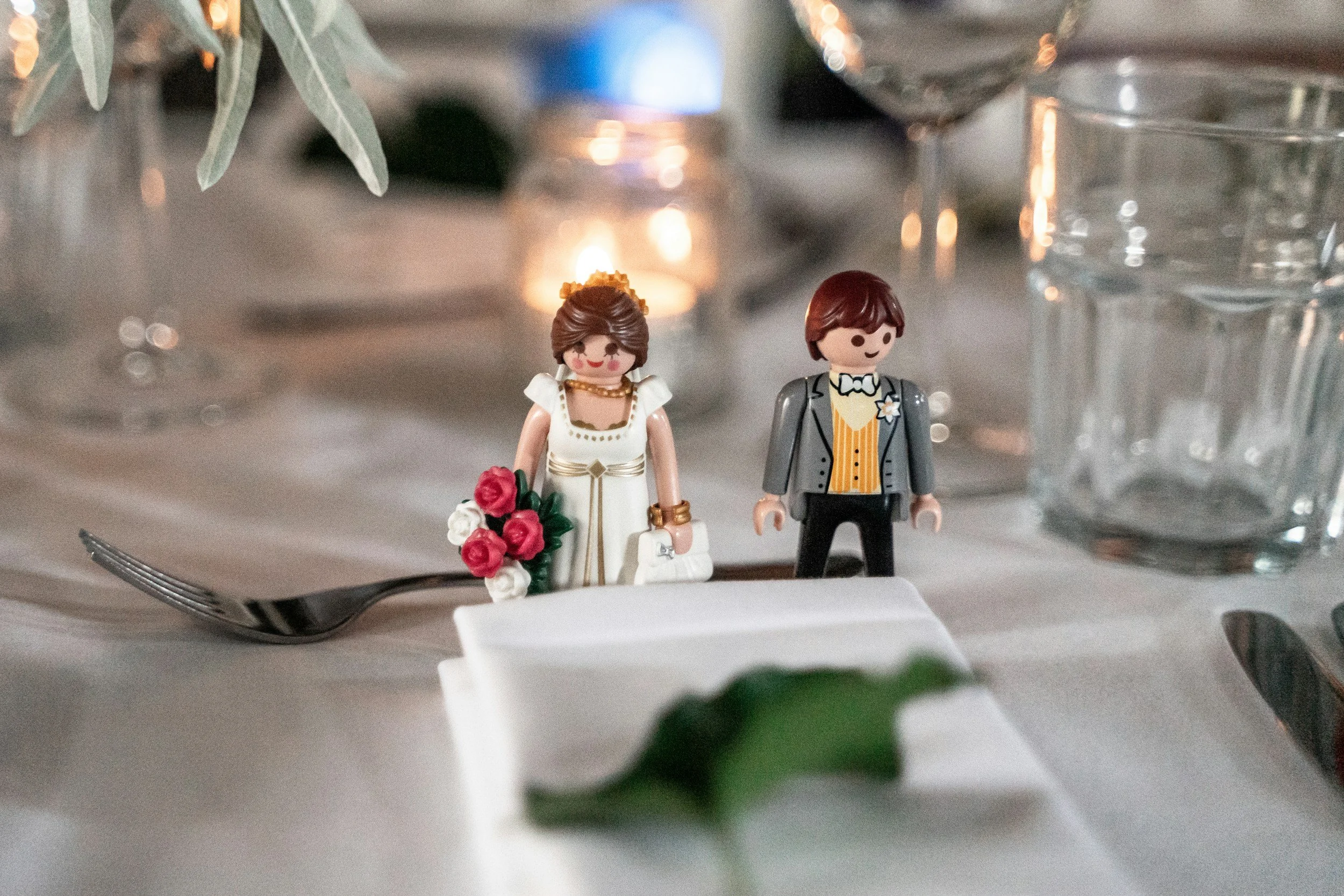 Close-up of small toy figurines of a bride and groom, with the bride holding a bouquet of red and white roses, set on a table with blurred glassware and greenery in the background.