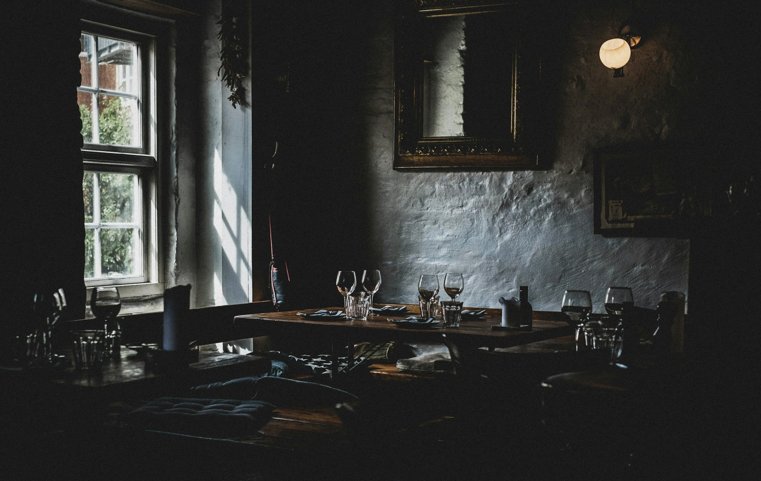 Dimly lit restaurant interior with a wooden table set for dinner, featuring wine glasses, water glasses, and black napkins. A window on the left lets in natural light, revealing a view of green foliage outside. The walls are textured and decorated with a framed mirror and a small wall-mounted lamp emitting a soft glow.