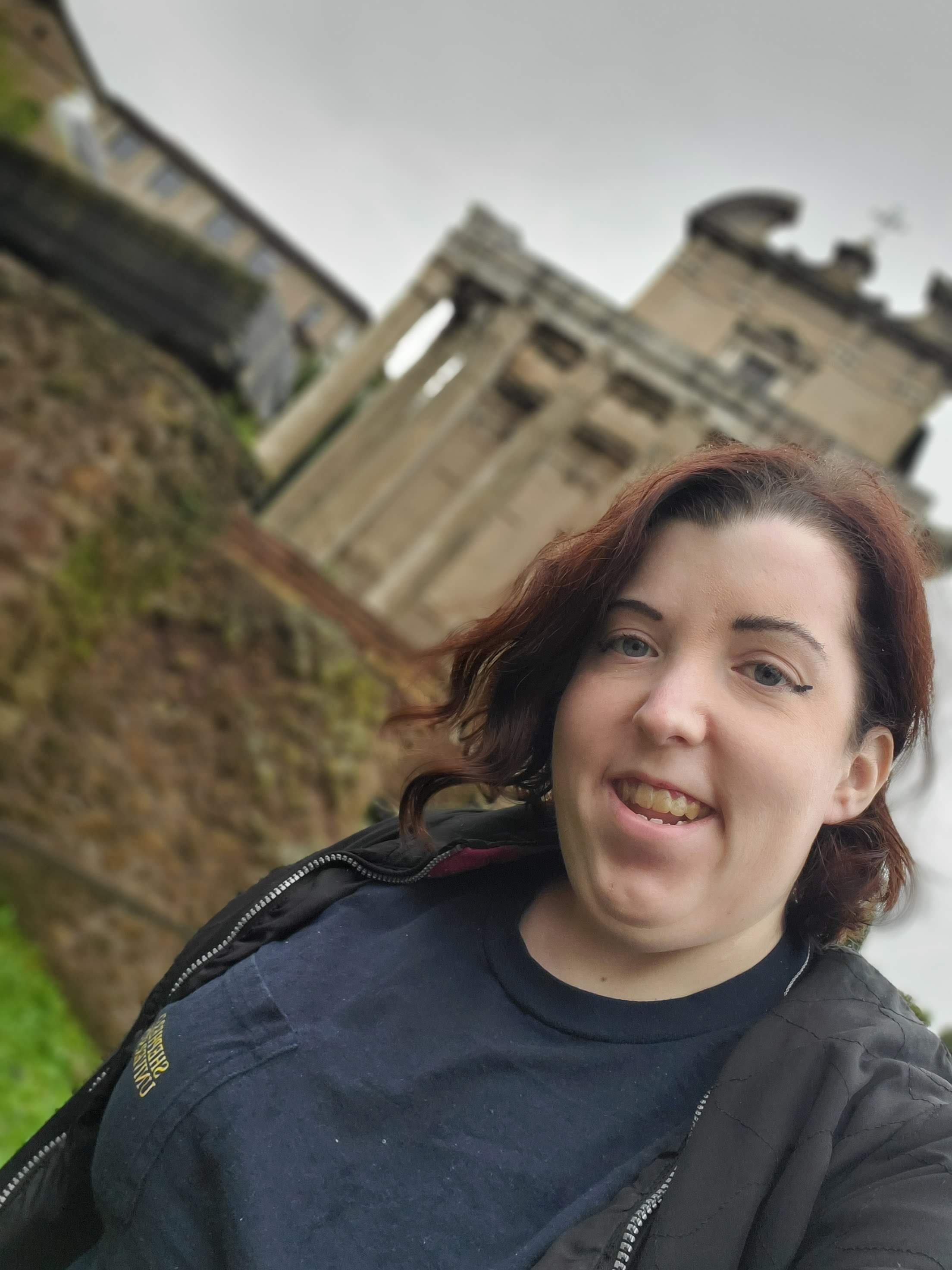 A woman with curly reddish-brown hair and blue eyes smiling outdoors near a historic stone archway and buildings.