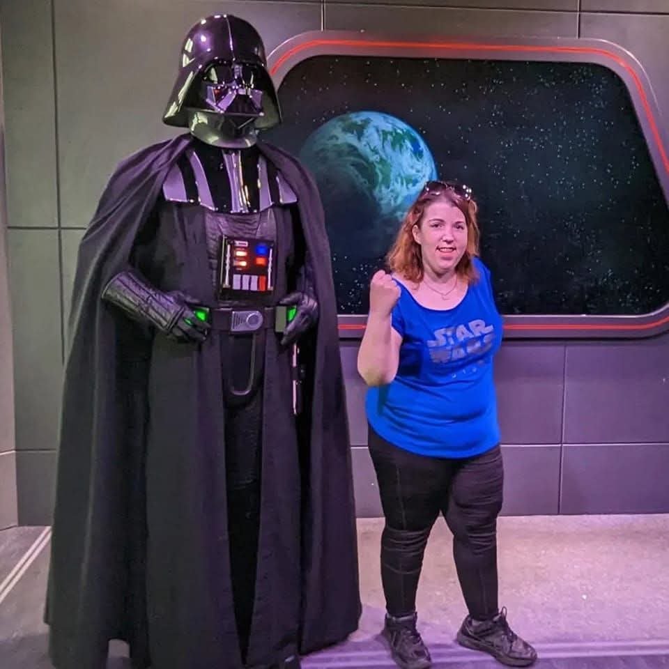 A woman in a blue Star Wars shirt posing with a life-sized Darth Vader figure inside a science fiction themed exhibit with a starry sky and planet Earth background.