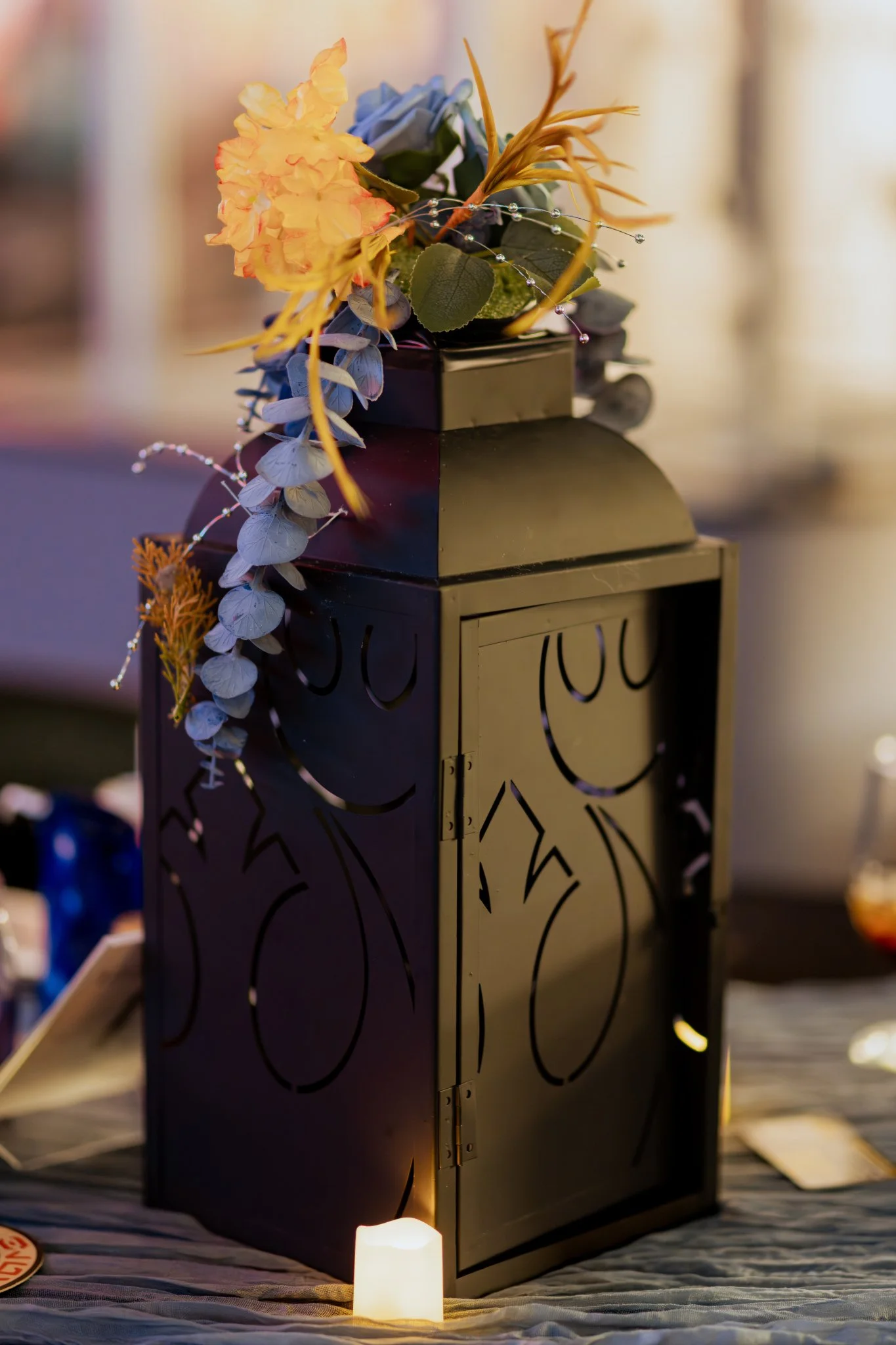 A decorative row of candles leading up to a black lantern with intricate cut-out designs, containing a floral arrangement with yellow, purple, and orange flowers, green leaves, and small decorative beads. The scene is softly lit with blurred background lighting.
