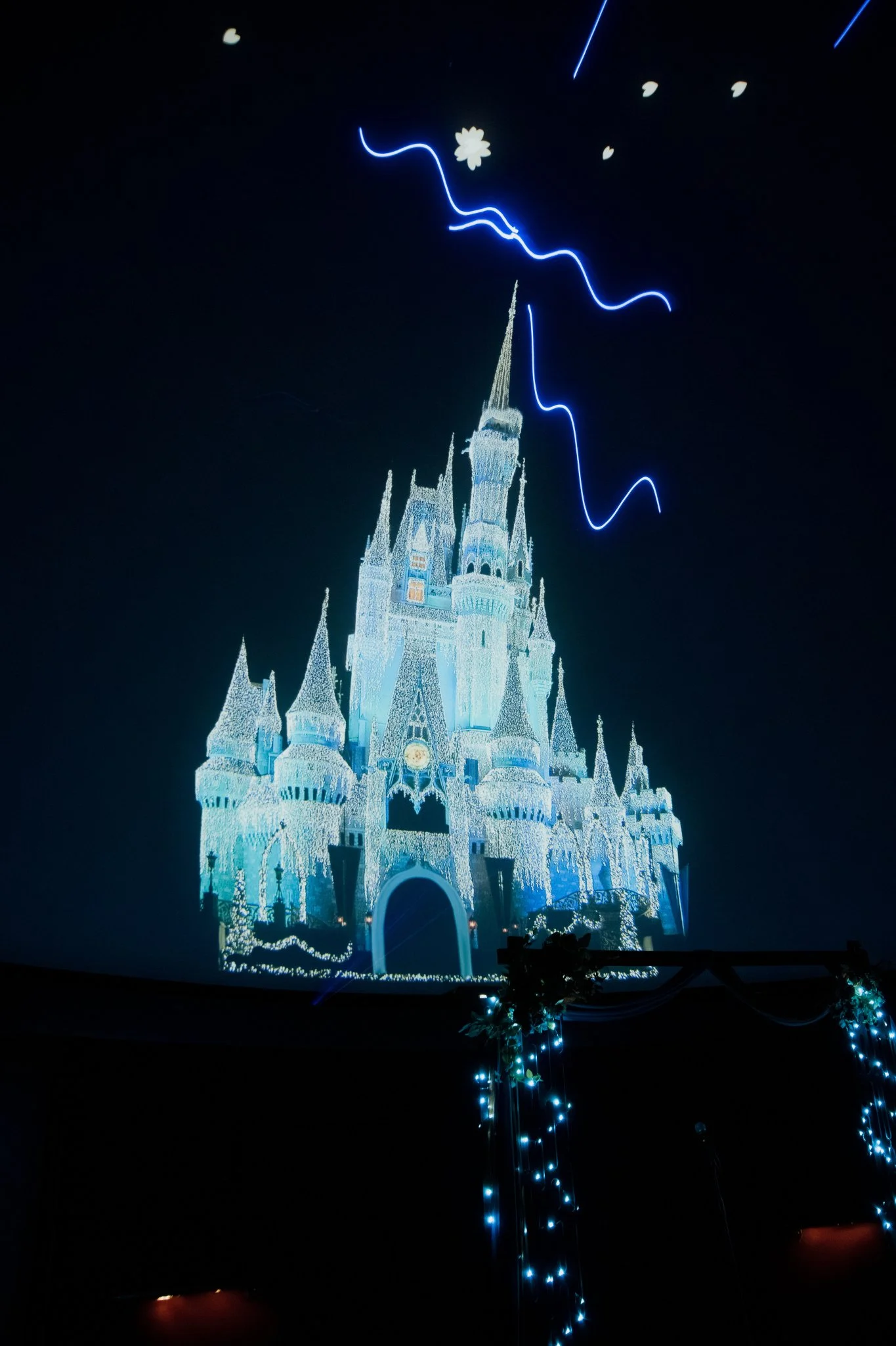 The image shows a castle illuminated with white and blue lights against a dark sky, with some blue light streaks and small decorative lights hanging in the foreground.