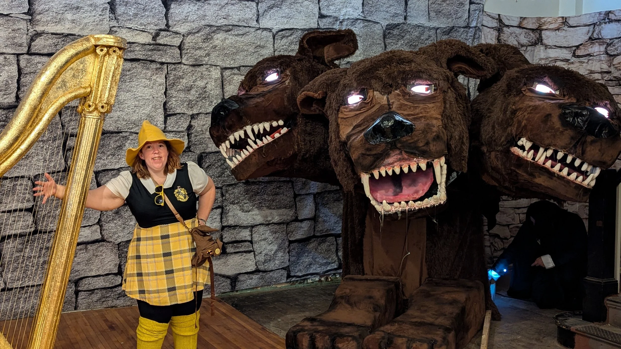 A woman dressed as a witch stands in front of large bear head sculptures with glowing eyes and open mouths, and a gold harp is partially visible on the left side, all set against a gray stone wall.