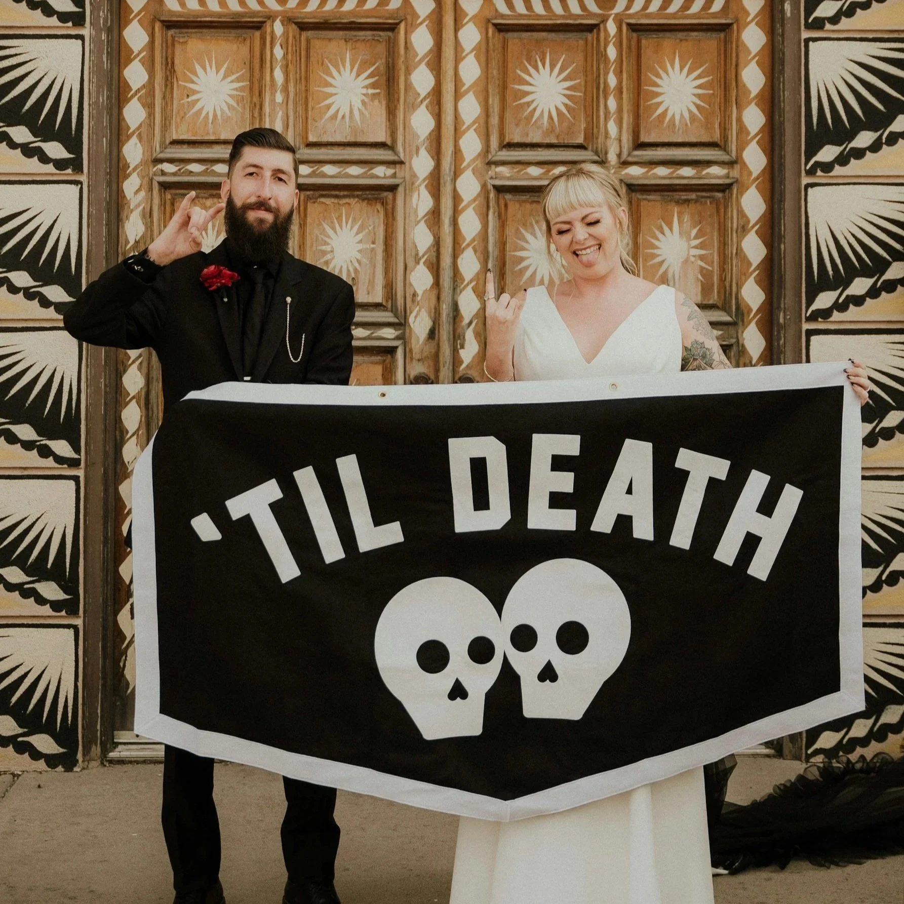 Two people standing behind a large sign with skull images, one man with a beard making a hand gesture and wearing a black suit, and one woman with blonde hair making a tongue out and middle finger gesture, both smiling and holding the sign.