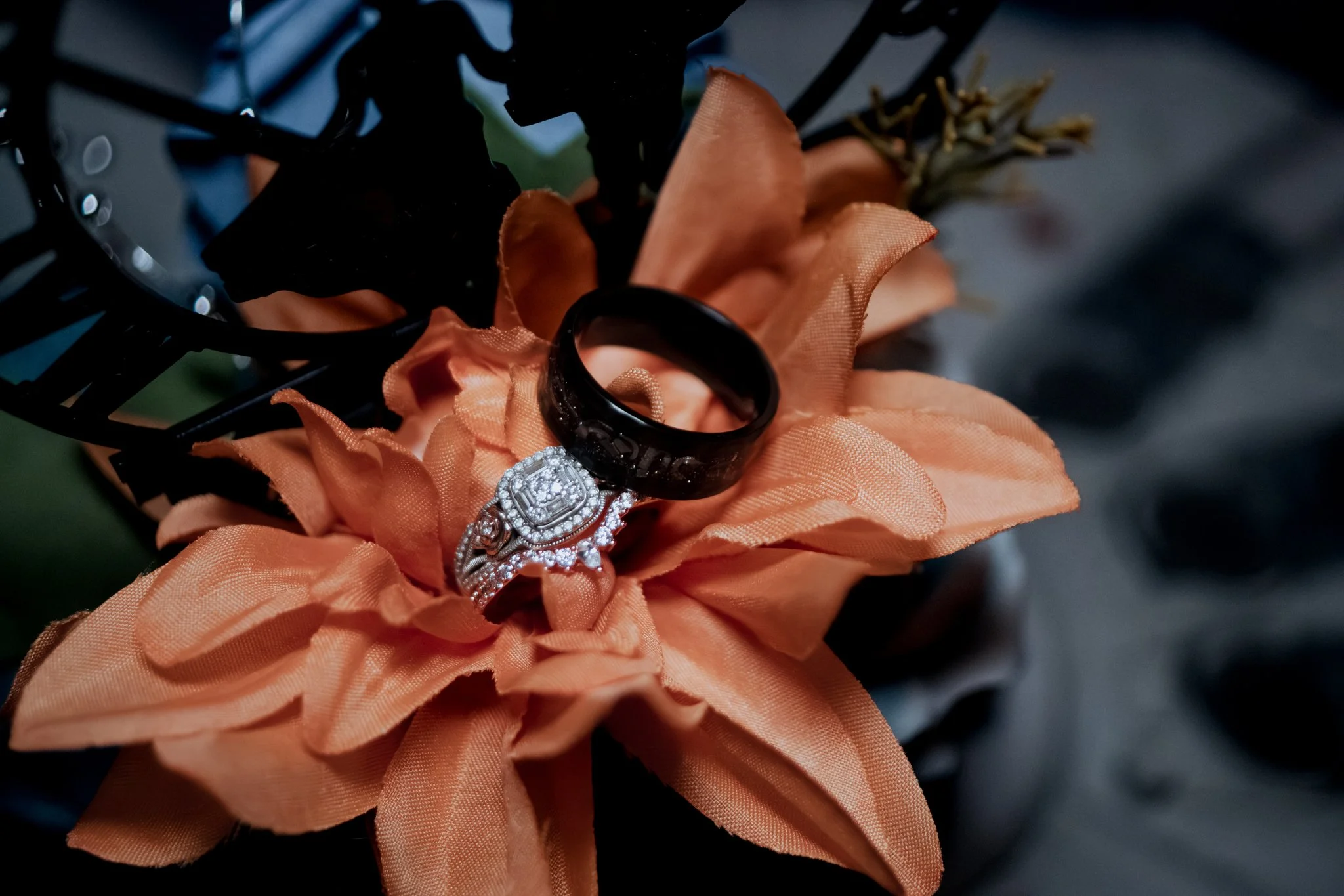 A close-up of a peach-colored artificial flower with a black ring and a diamond engagement ring resting on its petals.
