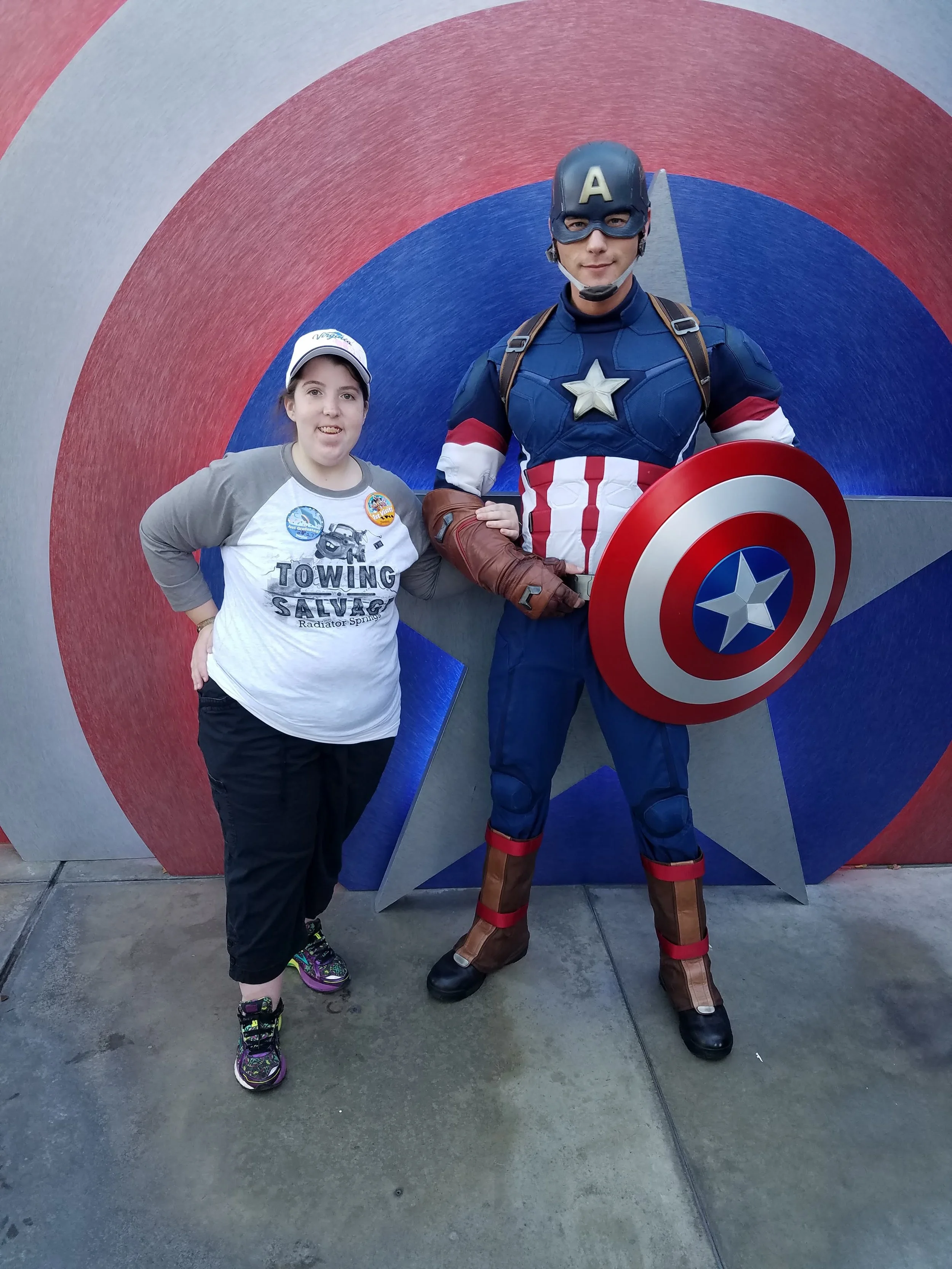 A young girl standing next to a man dressed as Captain America in front of a blue, red, and gray target background. The girl is wearing a white and gray T-shirt, black pants, colorful sneakers, and a white cap. The man is wearing a Captain America costume with a shield, helmet with an 'A', and brown boots.