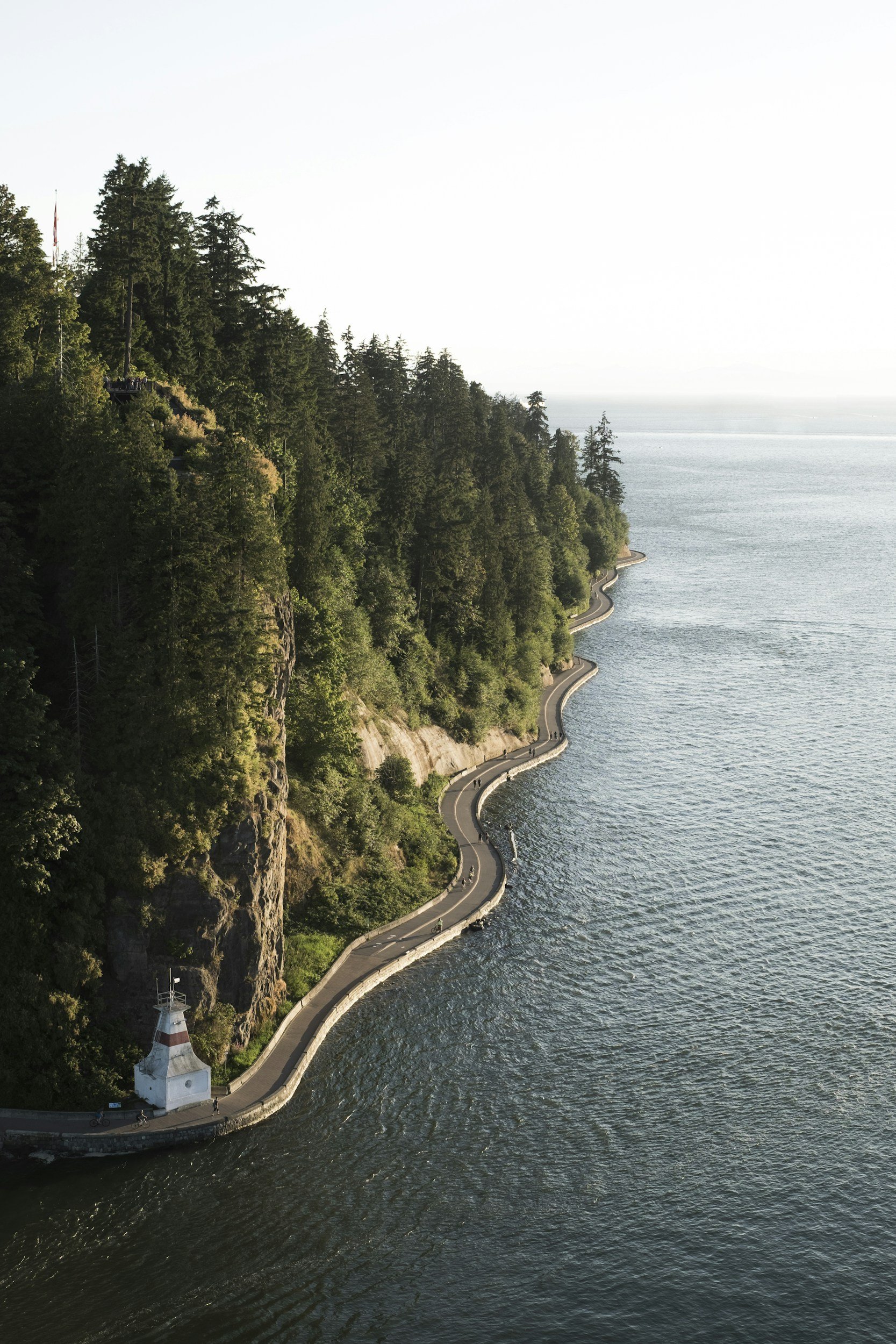 A winding road runs along the edge of a body of water, with lush green trees on a steep hillside and a small white lighthouse at the bottom.
