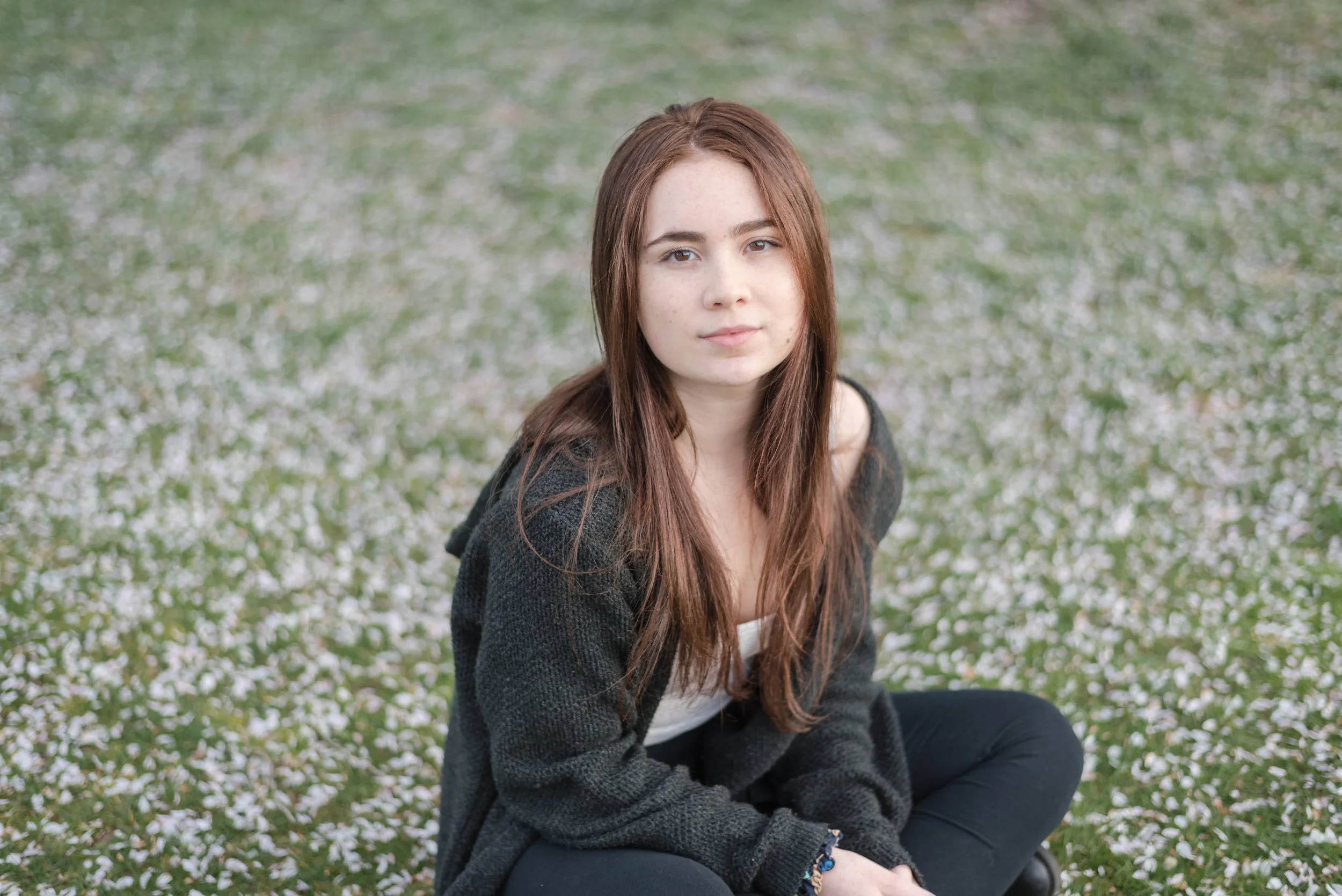 A young woman with long brown hair, sitting cross-legged outdoors on a field covered in small white flowers, looking at the camera.
