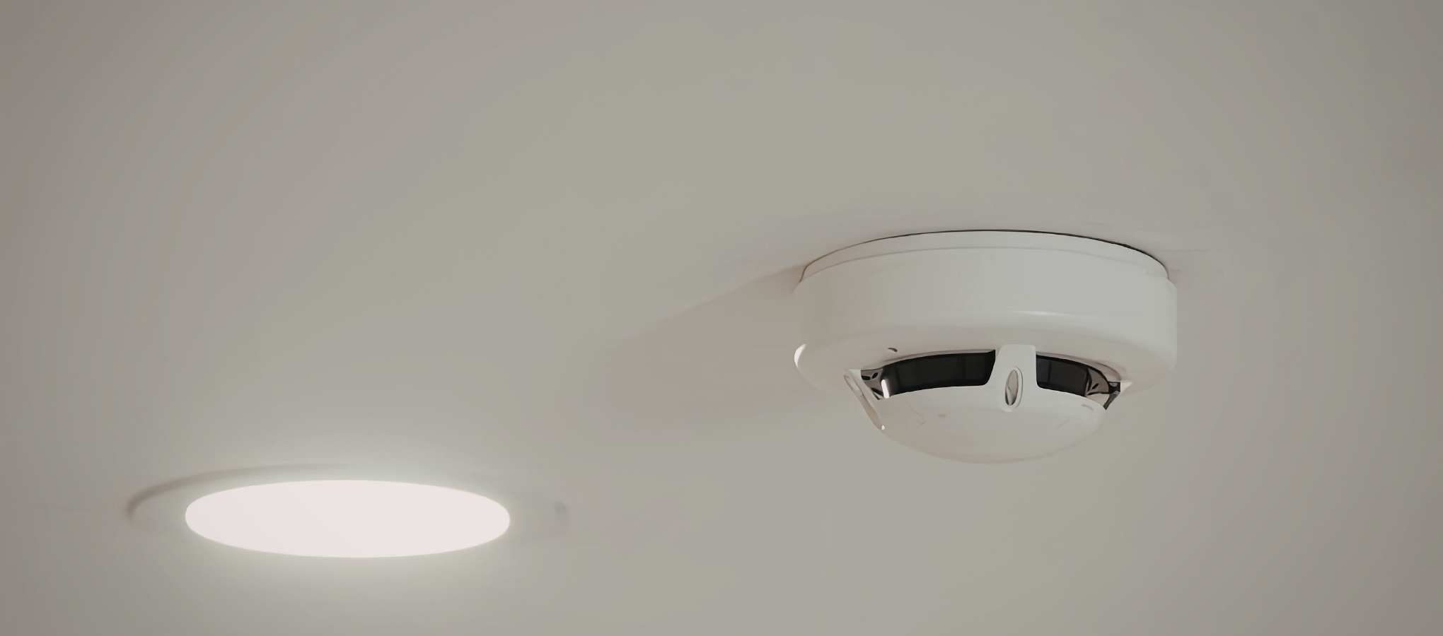 A ceiling-mounted smoke detector with a circular design and black vents, next to a round, bright LED ceiling light.