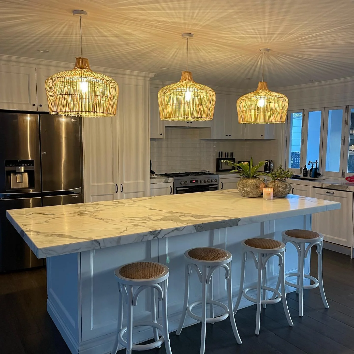 A modern kitchen with a large white marble island, three woven pendant lights, stainless steel refrigerator, white cabinets, and a window with a view outside.
