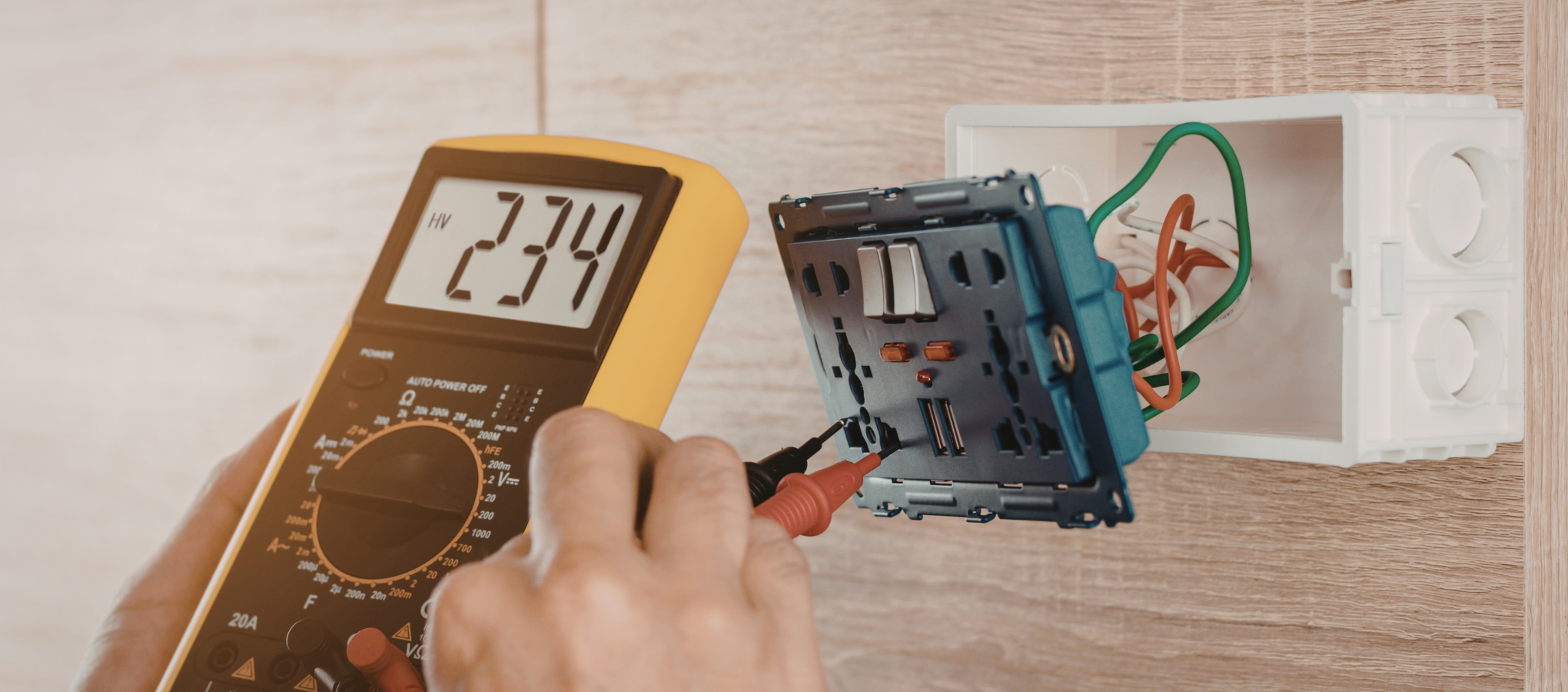 A person using a multimeter to check electrical wiring inside an electrical outlet box on a wall.