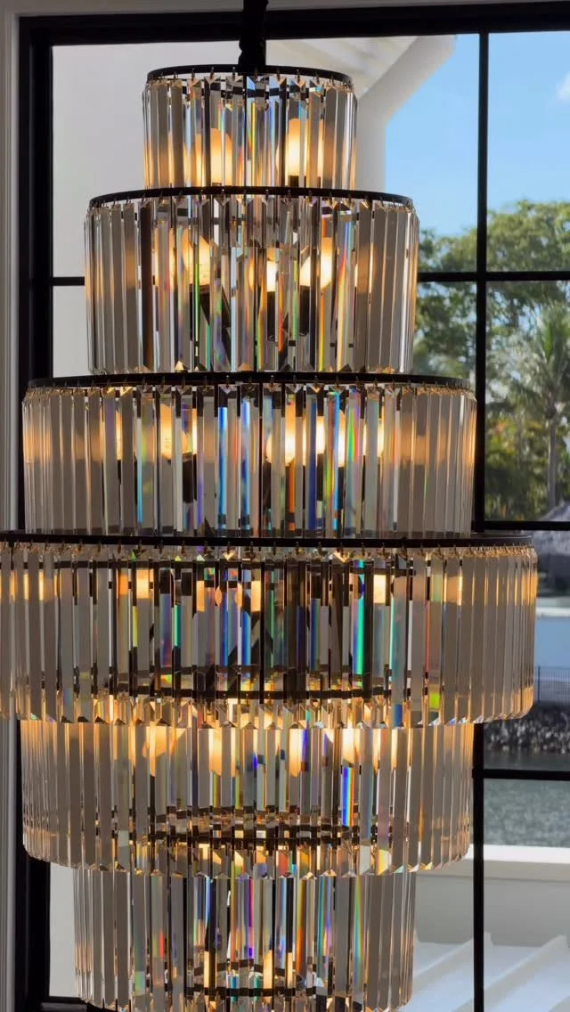Close-up of an elegant multi-tiered crystal chandelier hanging in front of window, with trees and blue sky visible outside.