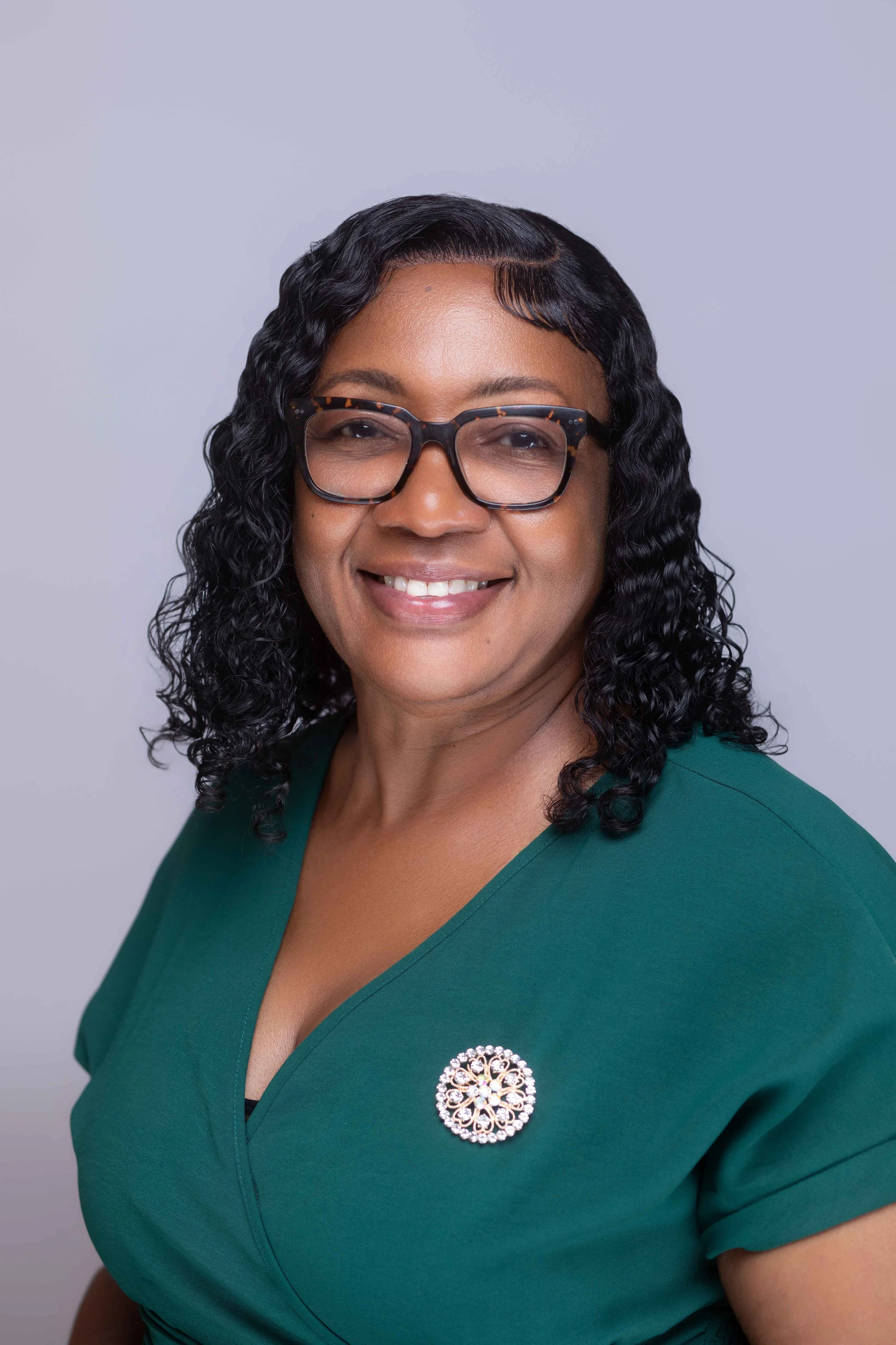 Close-up portrait of a smiling woman wearing black-rimmed glasses, a teal dress with a decorative brooch, and curly black hair, against a plain light gray background.