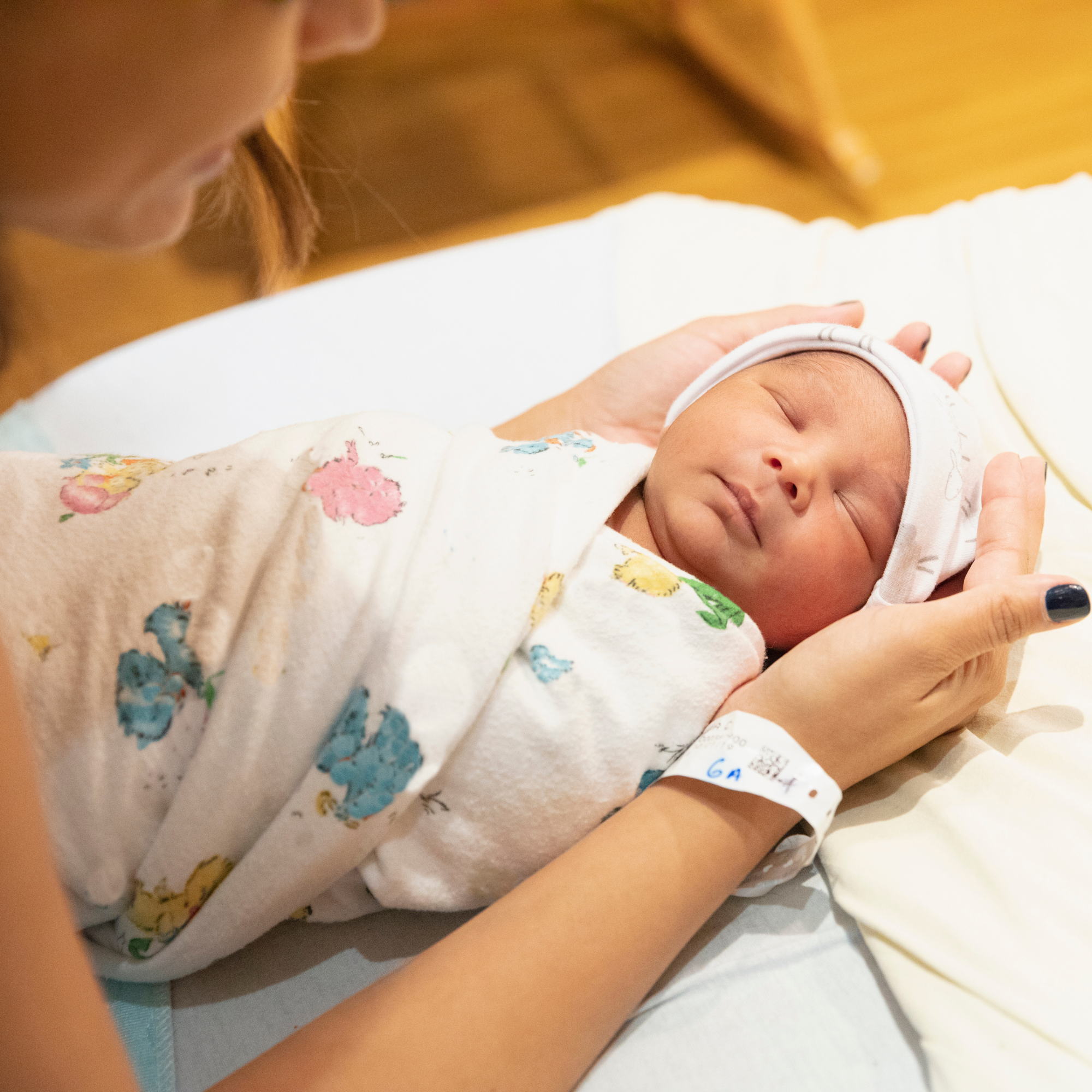 Close-up of a newborn baby sleeping peacefully, laying on a hospital bed, with a nurse gently supporting the baby's head. The baby is wearing a white hat and a blanket with colorful animal prints.
