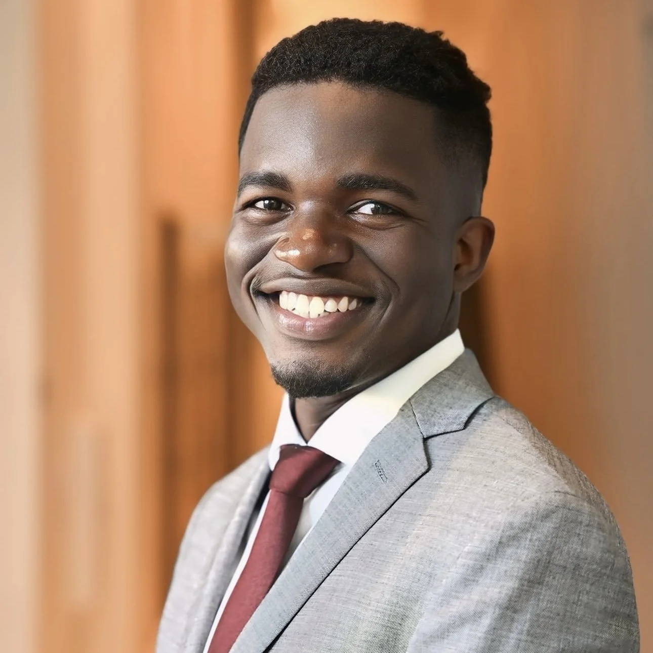 A young African American man in a gray suit and maroon tie, smiling with teeth showing, posing indoors with a wooden background.