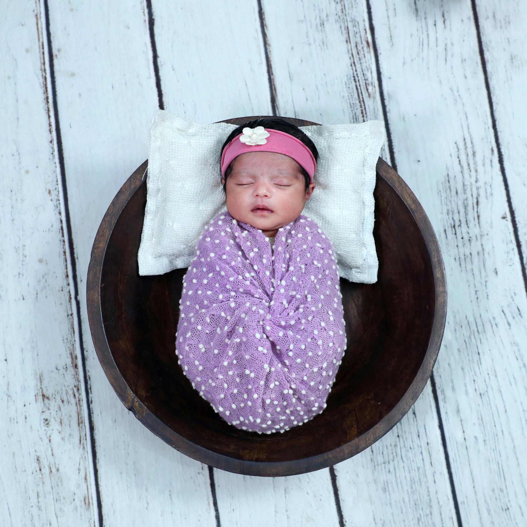 A sleeping baby girl with a pink headband with a white flower, lying on a white textured blanket in a wooden bed frame, against a white wooden plank background.