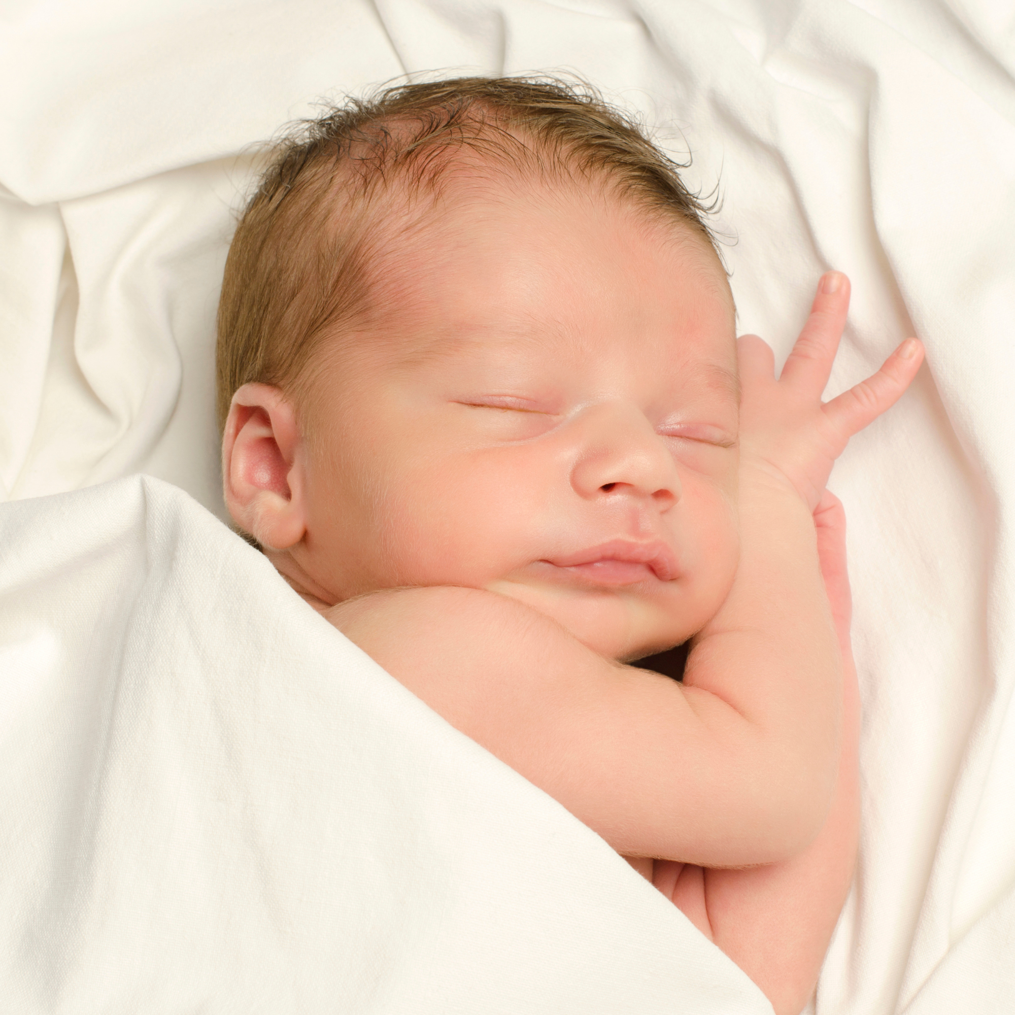 Close-up of a sleeping baby with light brown hair, lying on a white sheet, with a finger gently touching the sheet near the baby's face.