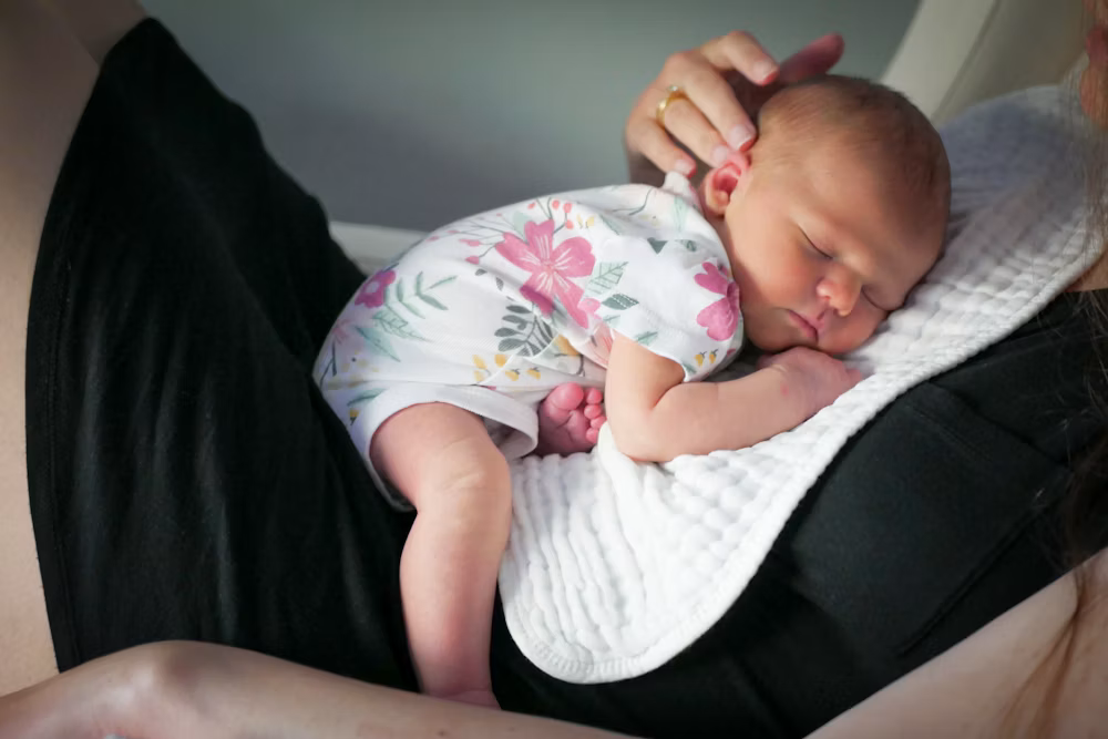 A baby sleeping on a woman's chest, resting on a white pillow, with the woman holding the baby's head gently.