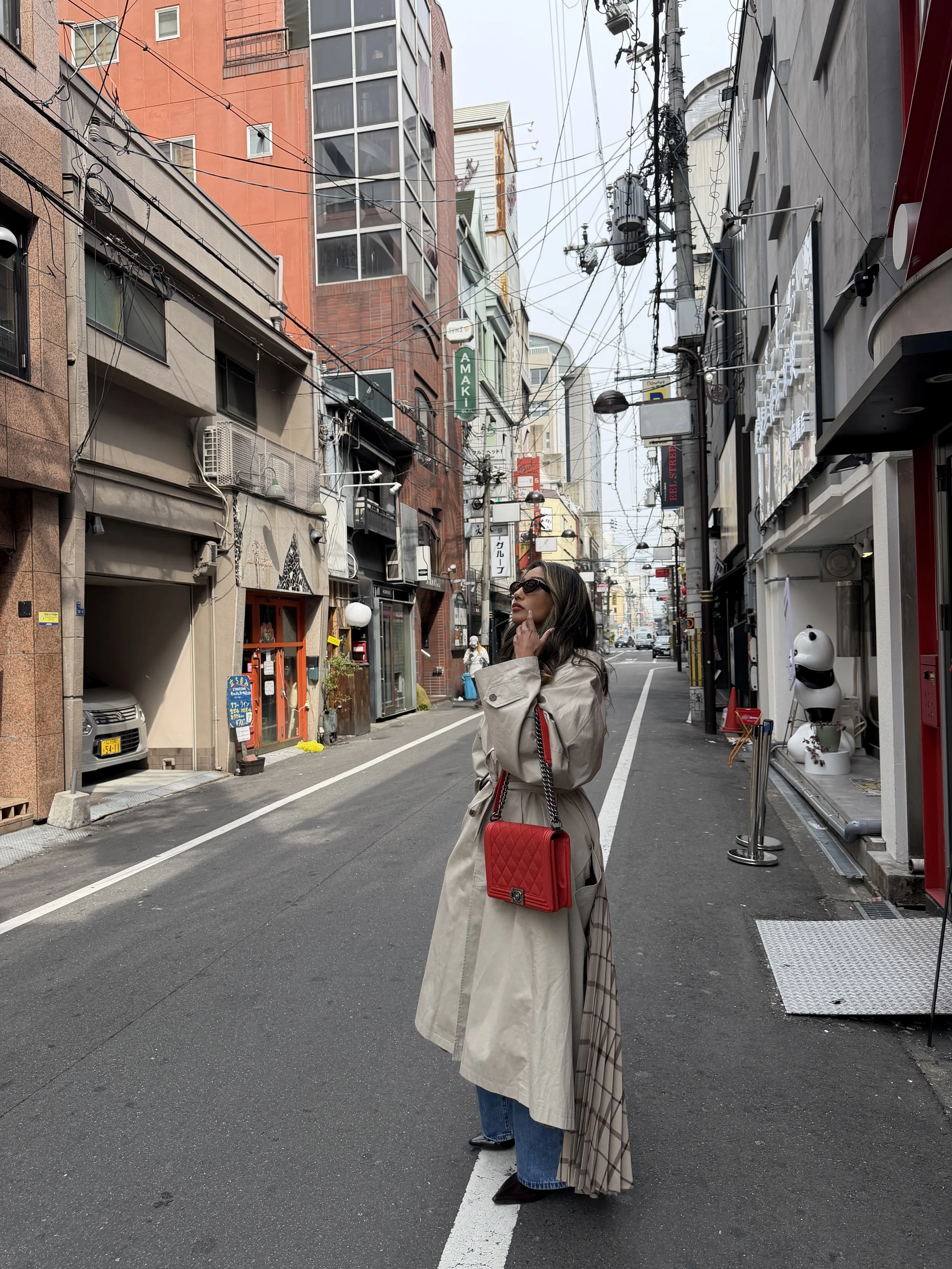 A woman in a Balenciaga beige trench coat and sunglasses stands on a city street with colorful buildings, power lines, and Japanese signs in the background. She carries a red quilted Chanel Boy Bag and looks to the side.