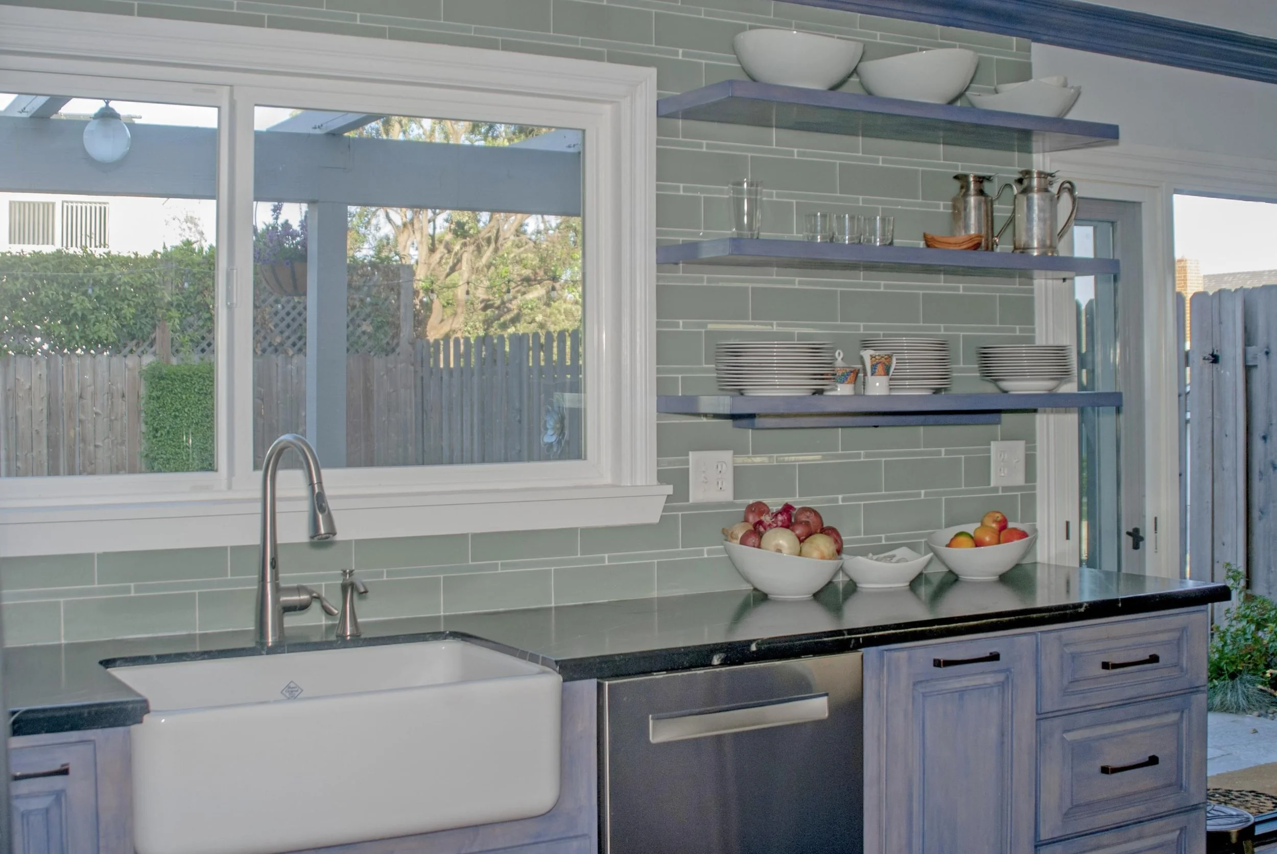 Kitchen with green subway tile backsplash, open shelves with bowls and glasses, a window showing outdoors, and bowls of apples and peaches on the counter.