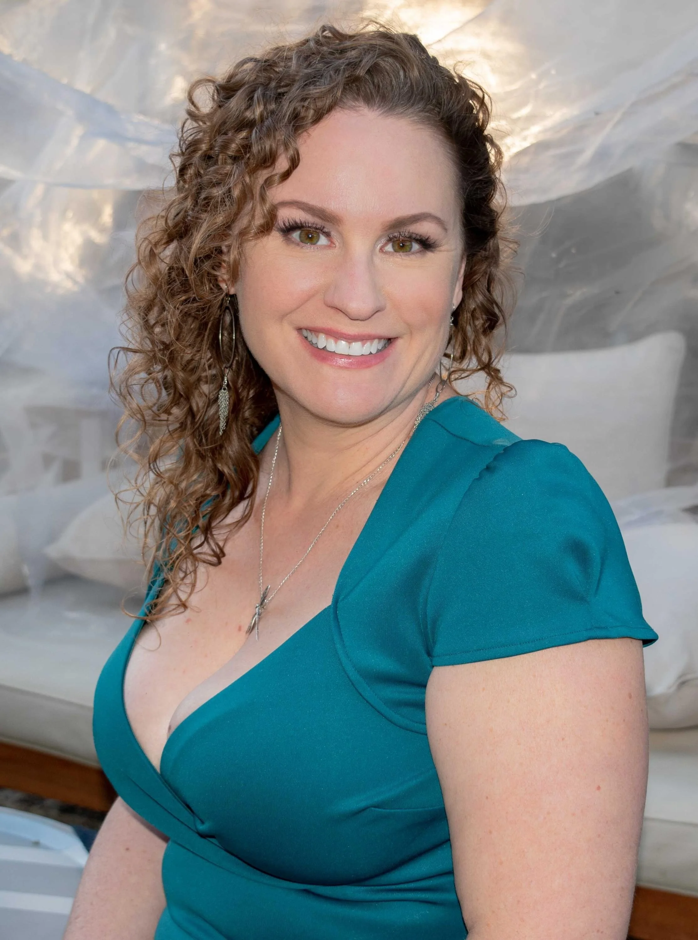 A woman with curly brown hair and hazel eyes smiling, wearing a teal dress and jewelry, in front of a blurred background that appears to be a bedroom with a bed and pillows.