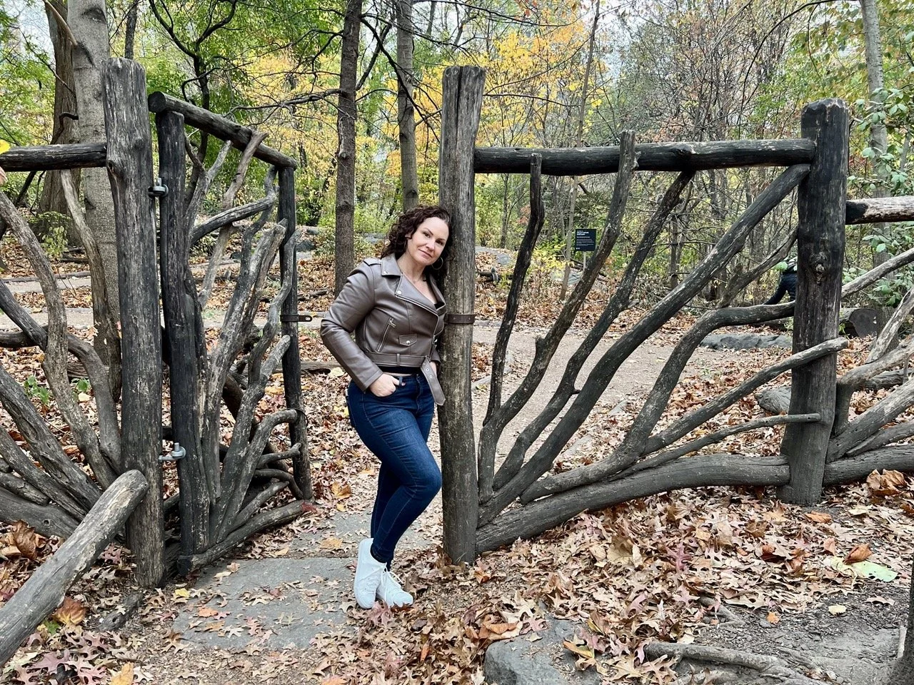 A woman with curly brown hair wearing a brown leather jacket, dark blue jeans, and white shoes standing next to a rustic wooden gate in a wooded park during autumn.
