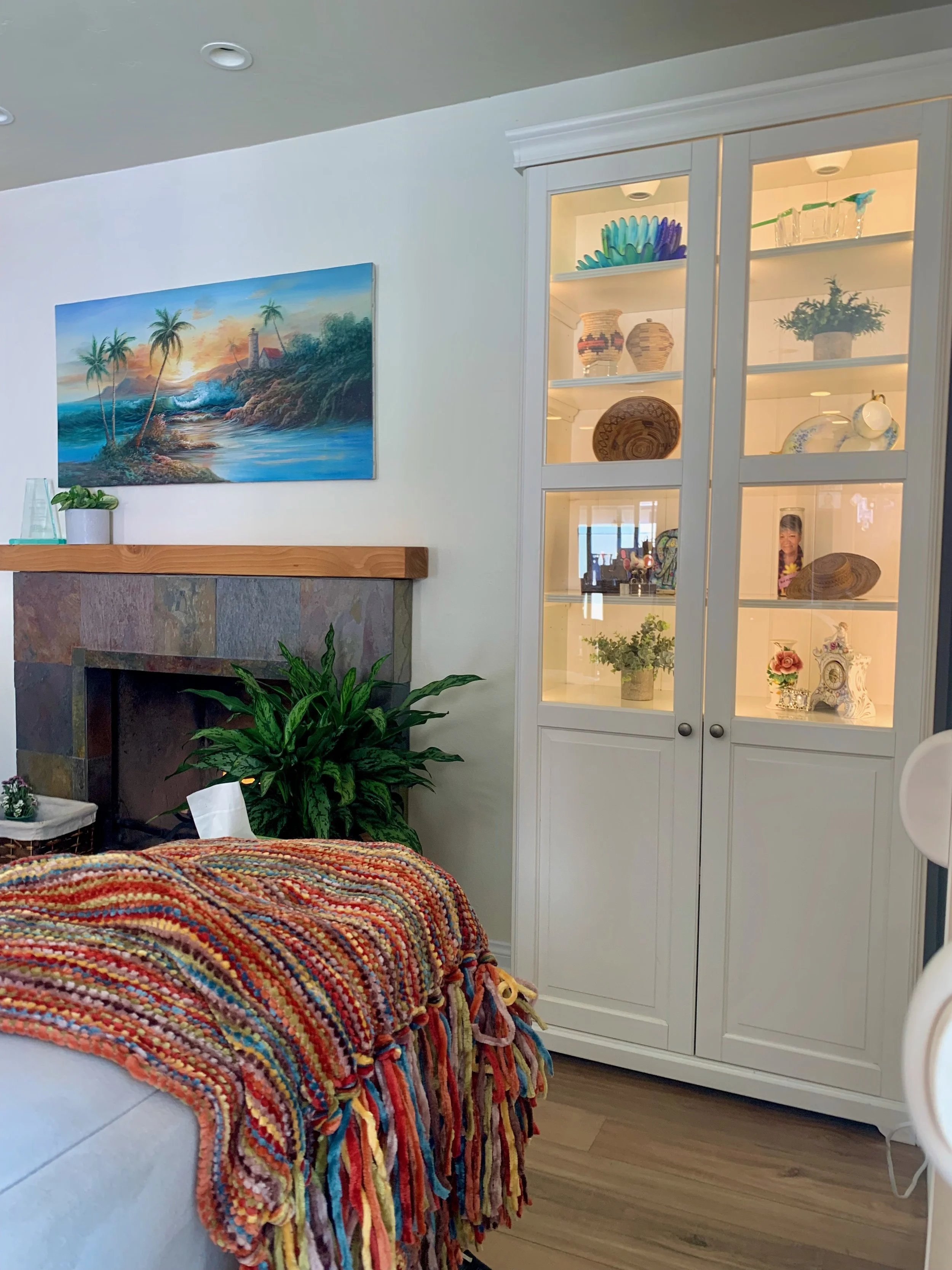 Living room corner with a bed covered by a multicolored knit blanket, a white display cabinet with decorative items, a painting of a tropical beach scene, and a wooden mantel with a plant.
