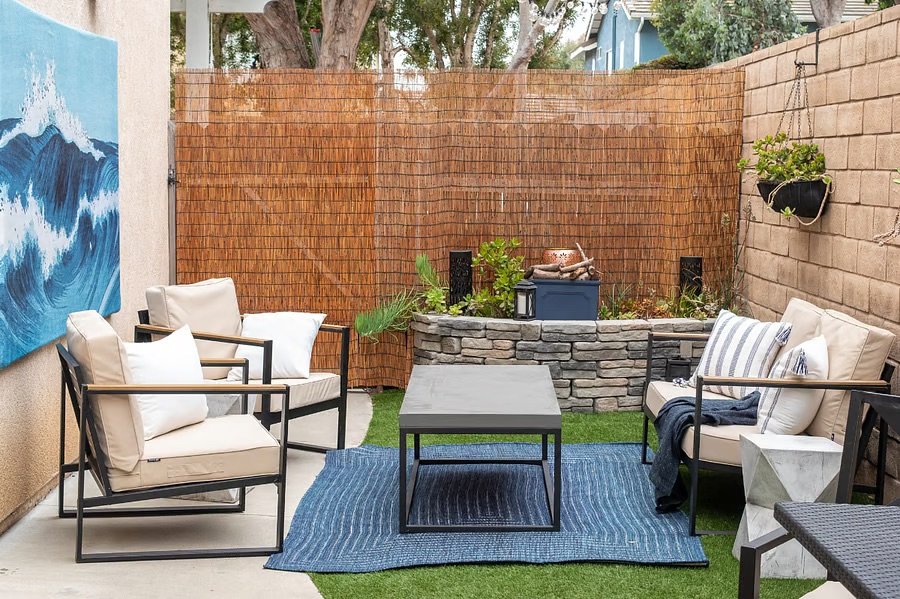 Backyard patio with seating area, a blue rug, beige cushions, a small coffee table, a stone fire pit, and a bamboo privacy screen.