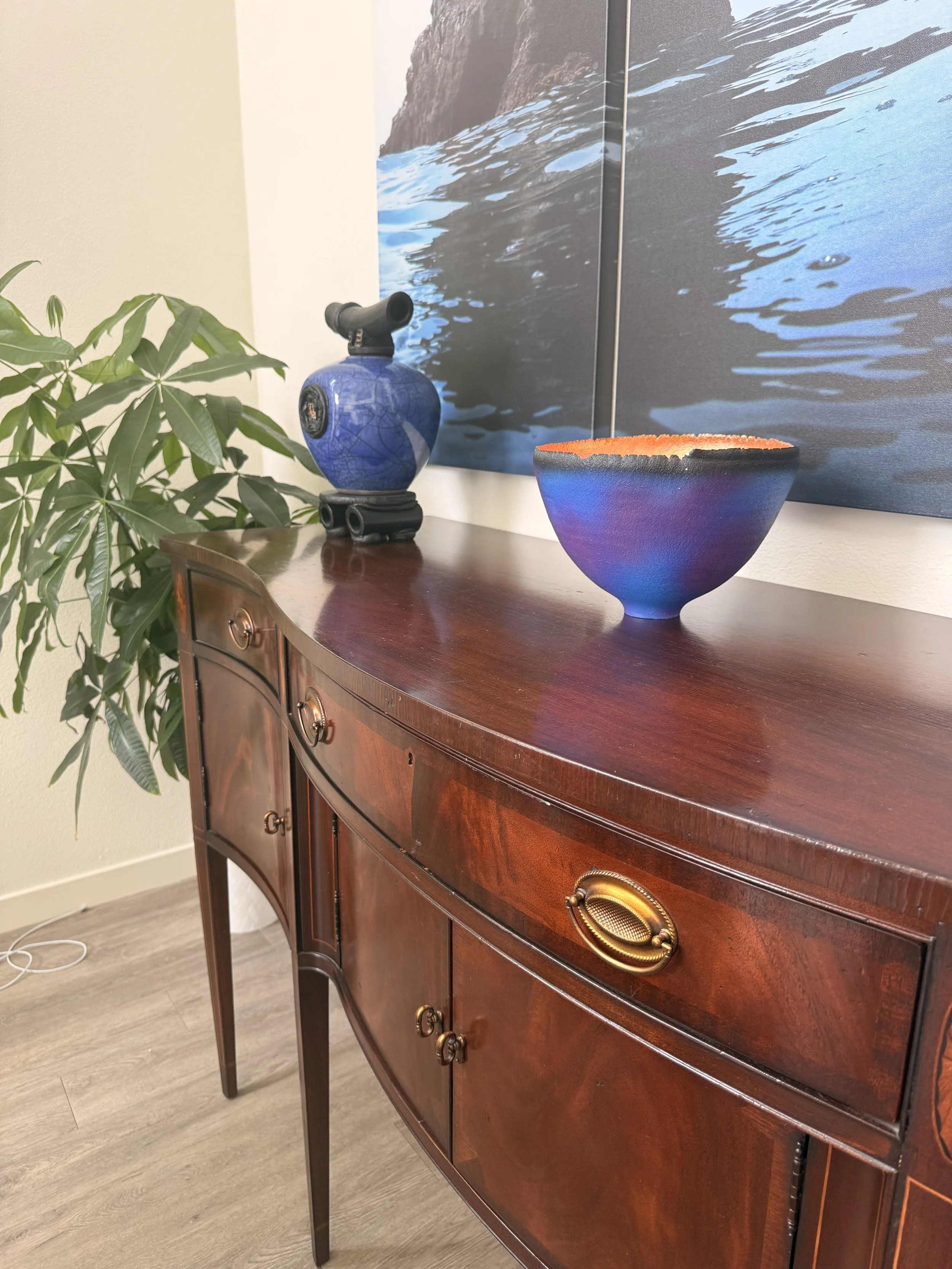 Decorative wooden sideboard with gold handles, topped with a ceramic vase, a multicolored bowl, and a large landscape photograph of water and rock formations, with a green plant on the side.