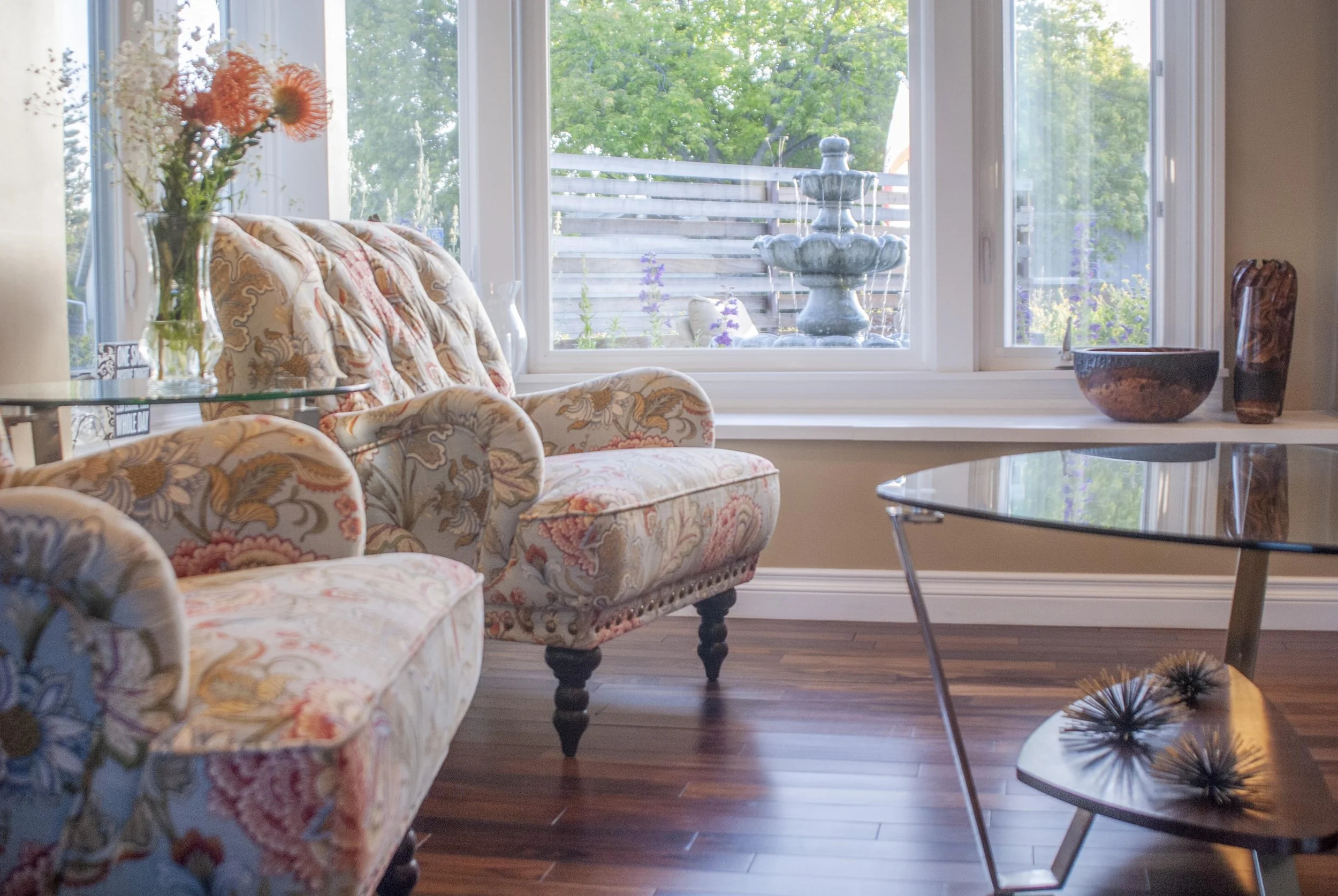 A sunlit living room with floral upholstered furniture, vases on a glass table, a window with a fountain outside, and a wooden floor.