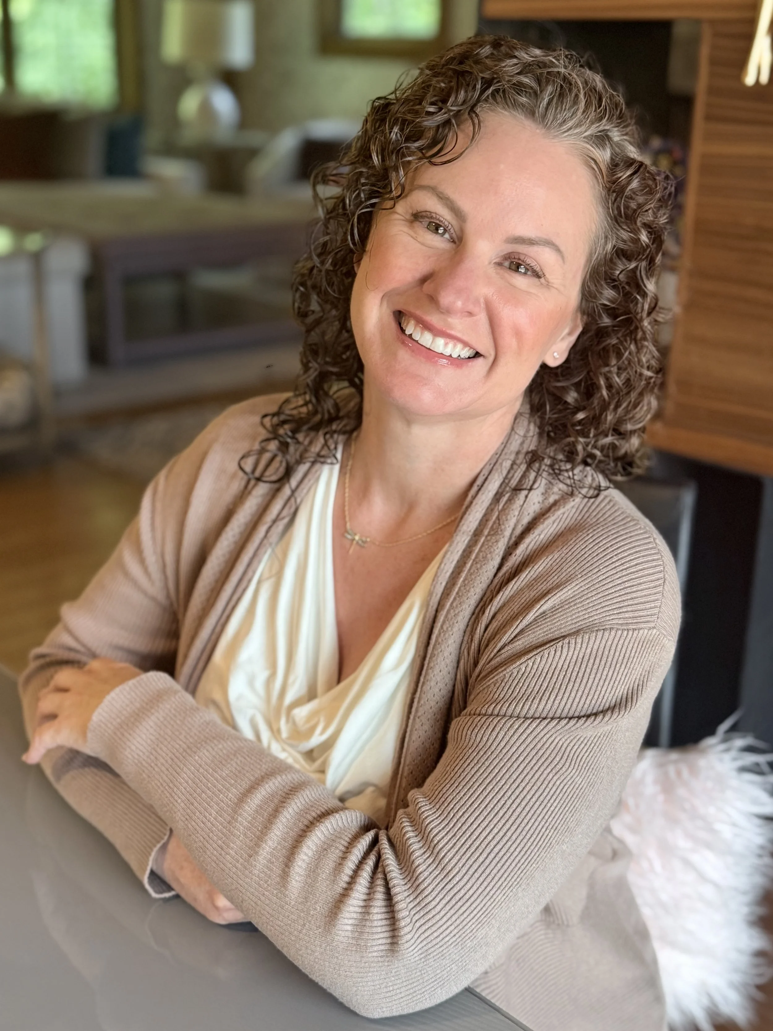 A smiling woman with curly hair sitting at a table with her arms crossed, in a cozy, well-lit room with windows and wooden furniture.