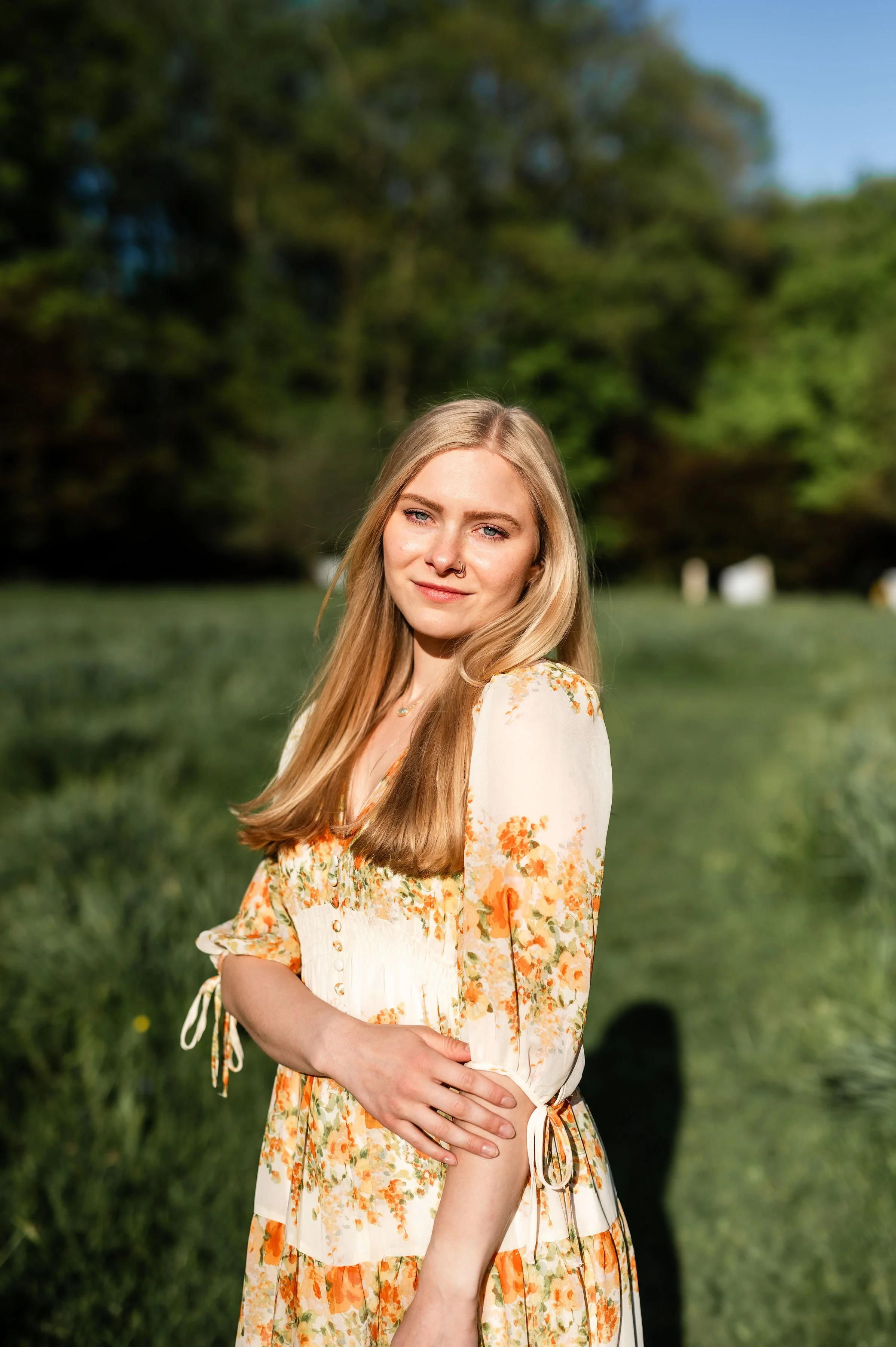 Young woman with blonde hair wearing a floral dress standing outdoors on a sunny day with trees and grass in the background.