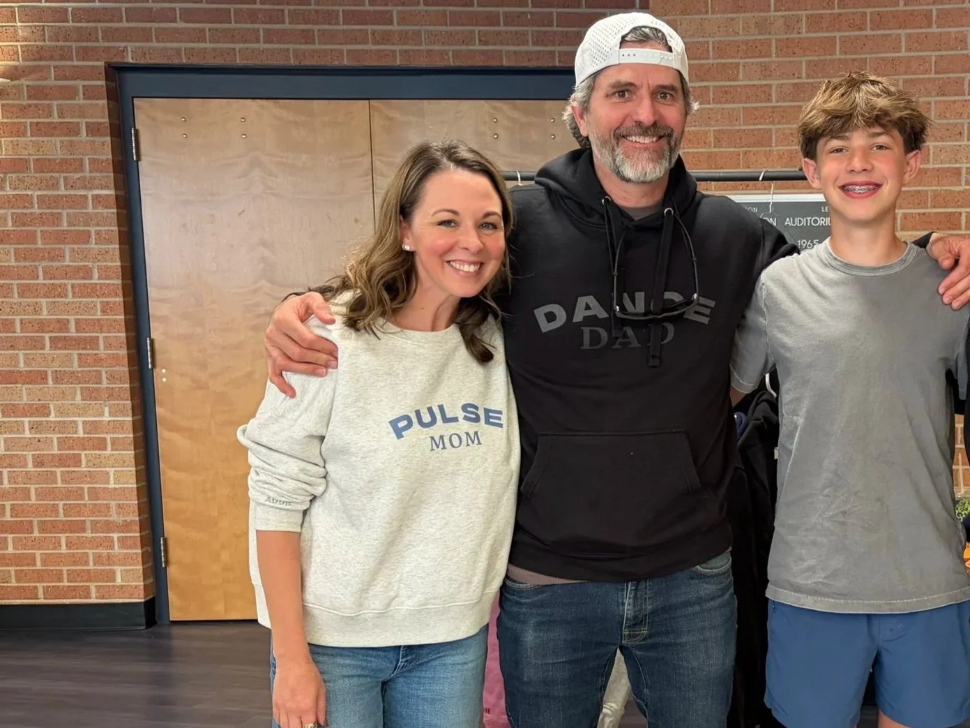 A smiling woman, man, and teenage boy standing together with their arms around each other indoors against a brick wall, all wearing casual clothing.