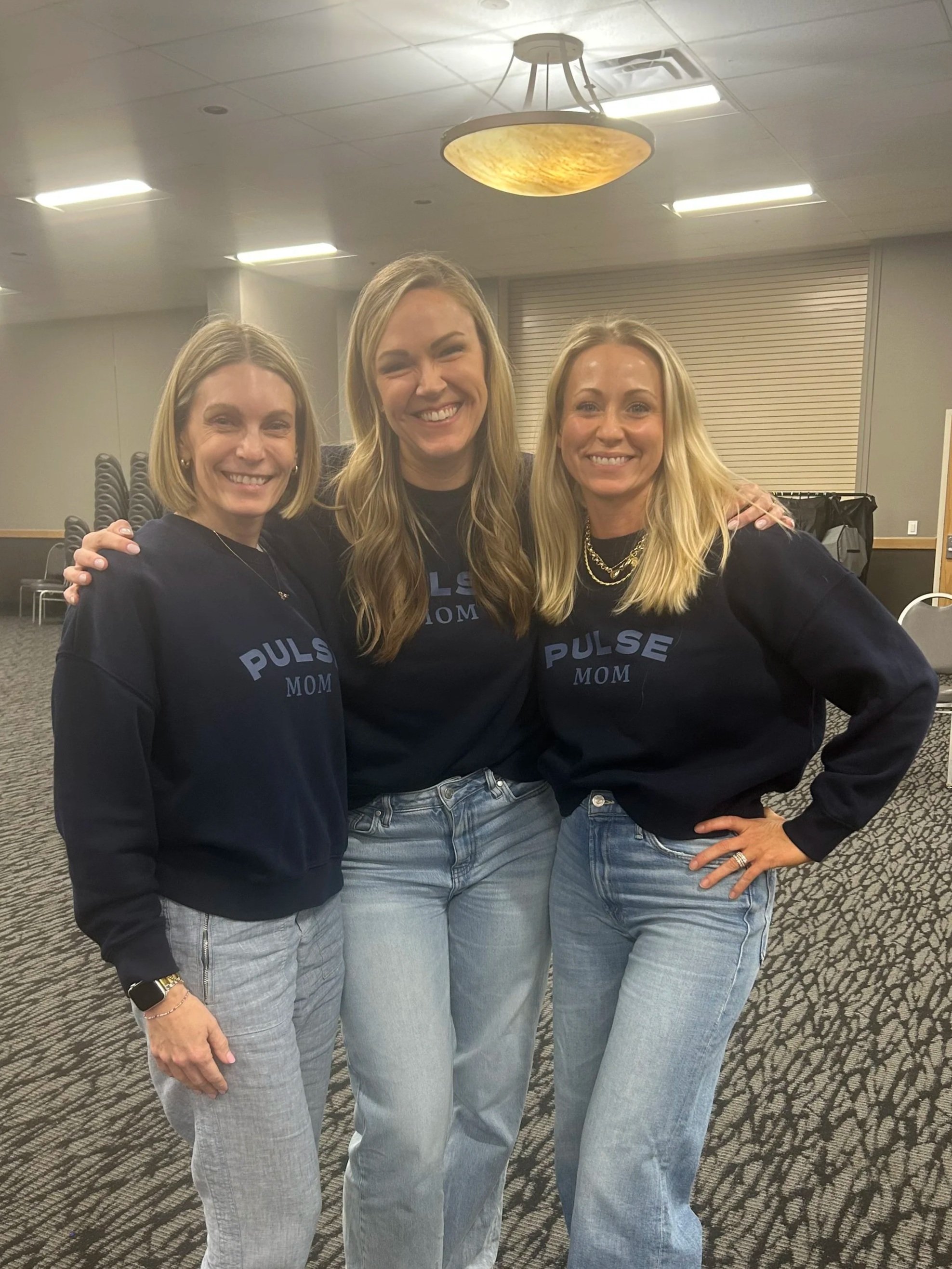 Three women standing close together and smiling for a photo in a room with chairs and a patterned carpet, all wearing black sweatshirts with blue text that says 'PULSE MOM'.