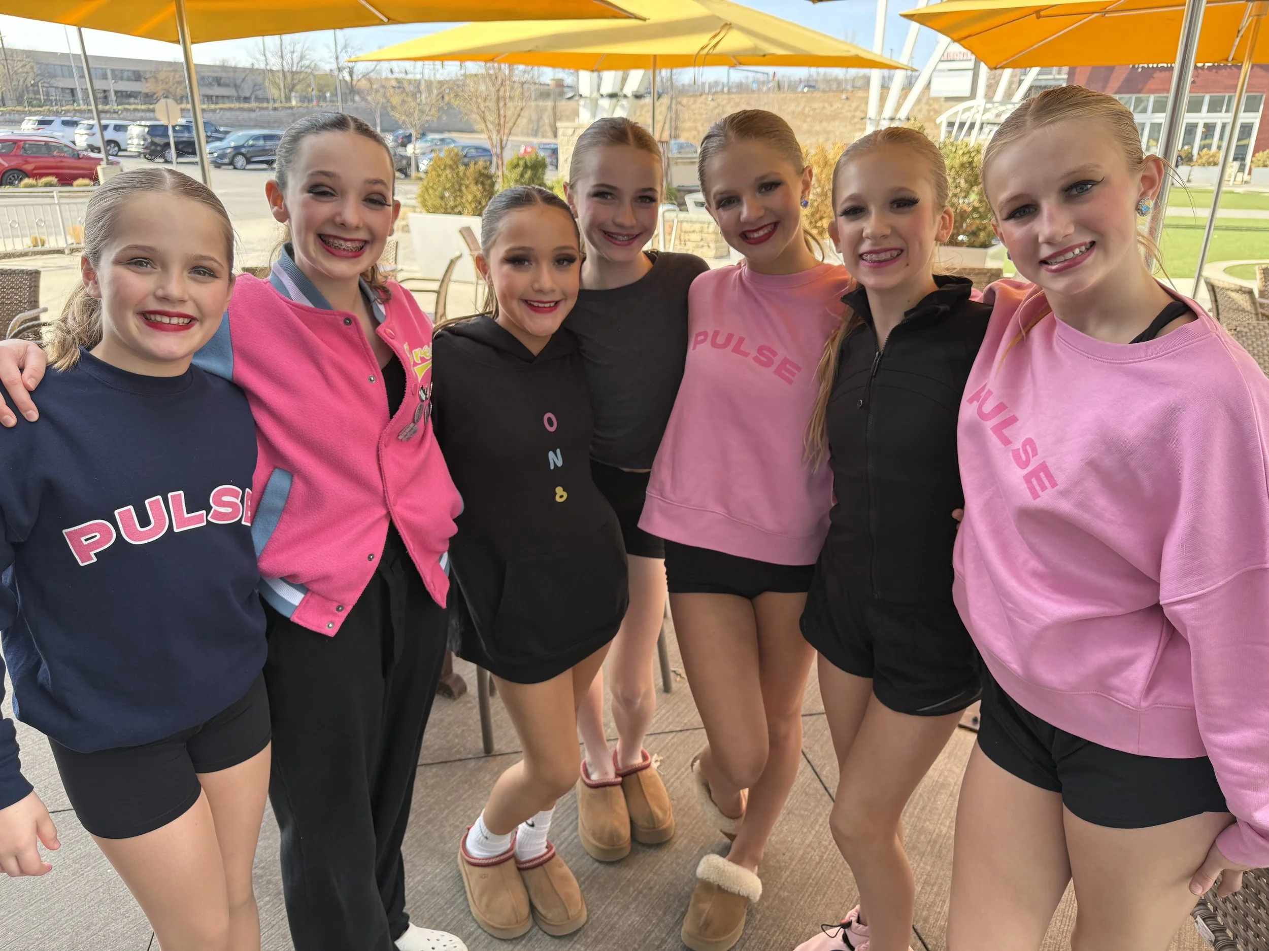 Group of young girls in casual and sporty outfits smiling and standing close together on a patio with outdoor seating and umbrellas.