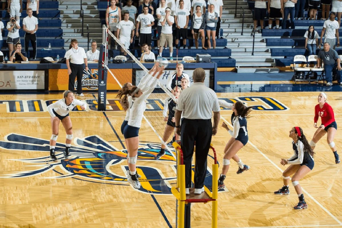 Women's volleyball game with players jumping to hit the ball over the net, spectators in the background, and an official overseeing the game.