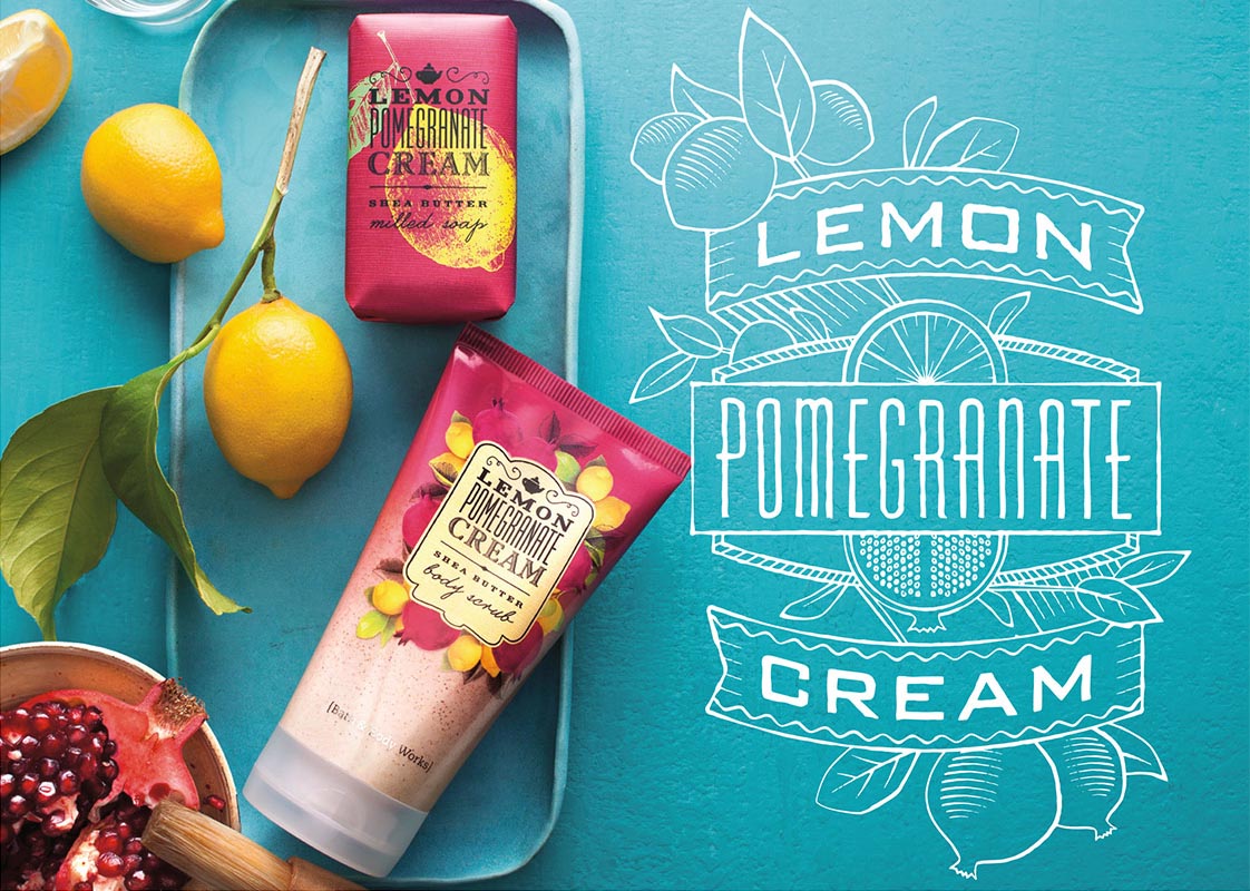 A bright blue tray with two yellow lemons and two packages of Lemon Pomegranate Cream Shea Butter body products; one is soap, and the other is body souffle. A pomegranate with seeds and a wooden spoon is partially visible at the bottom left corner.