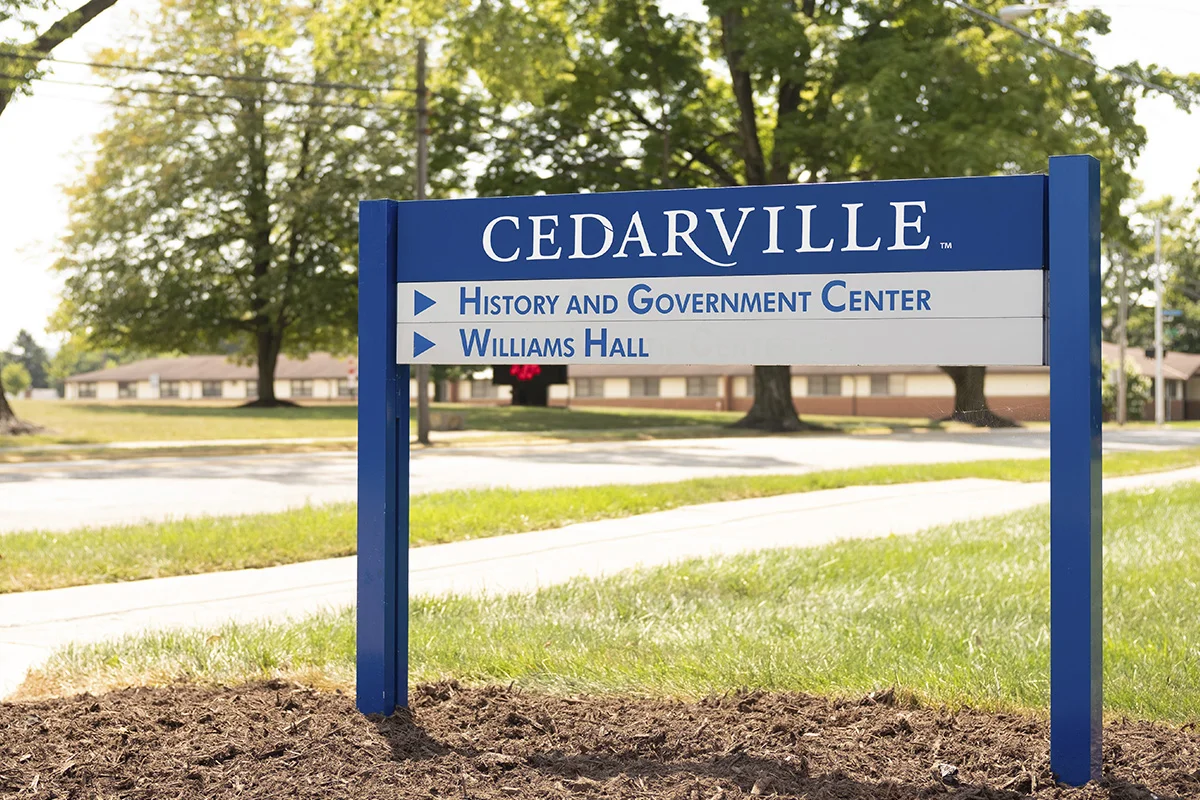 A blue and white sign for Cedarville town indicating directions to the History and Government Center and Williams Hall, with trees and a building in the background.
