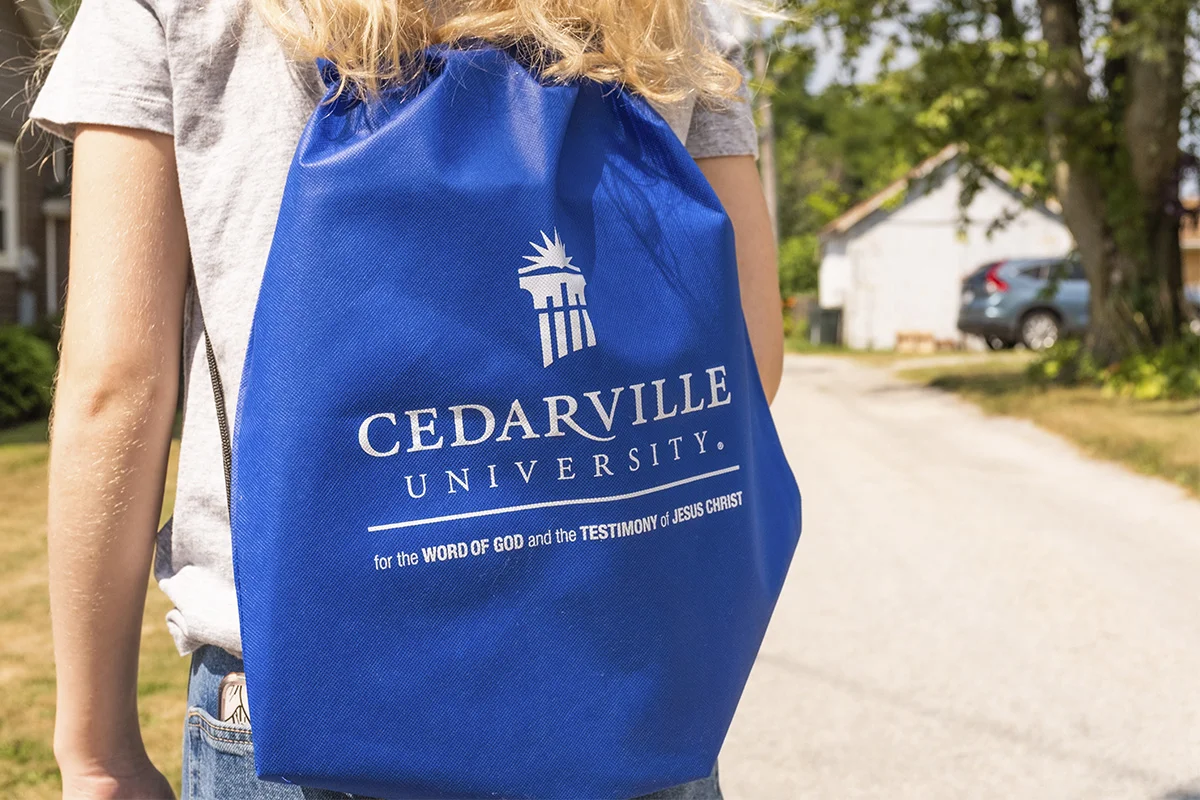 Person wearing a gray T-shirt and jeans carrying a blue Cedarville University drawstring backpack outdoors on a sunny day, with trees and houses in the background.
