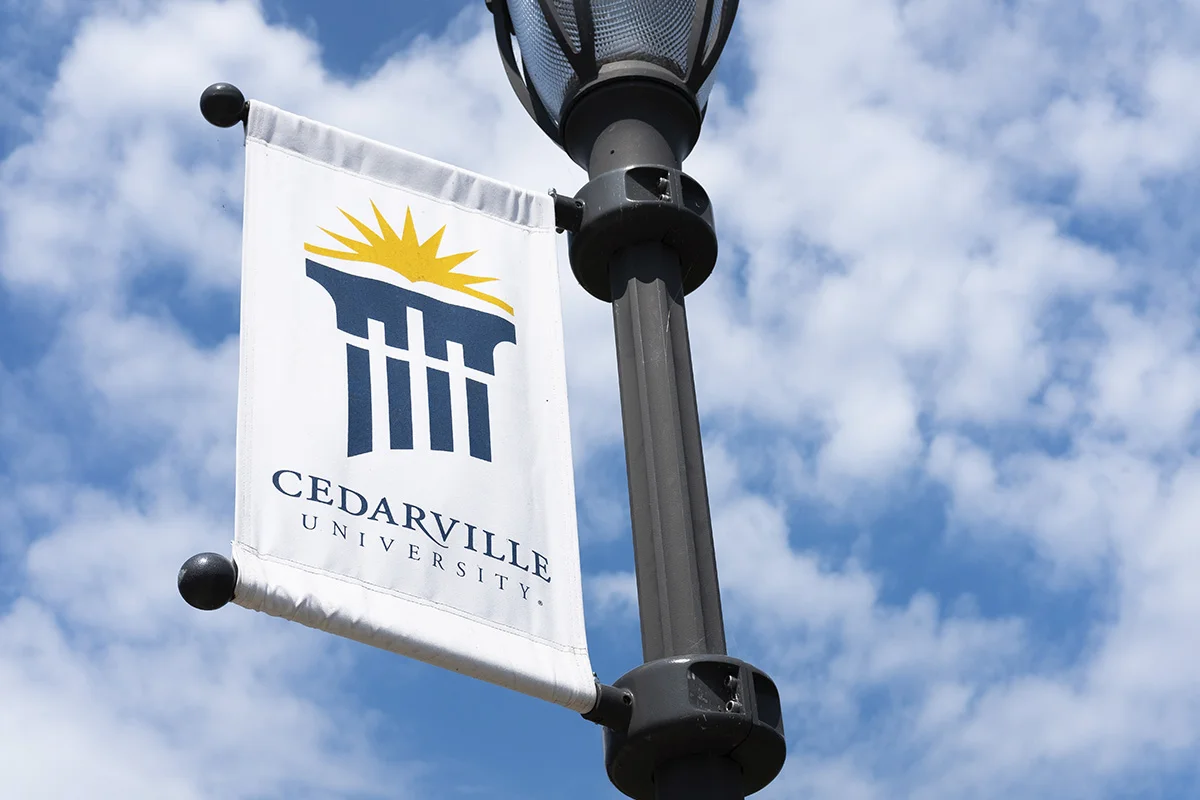 Cedarville University banner hanging from a street lamp against a partly cloudy sky.