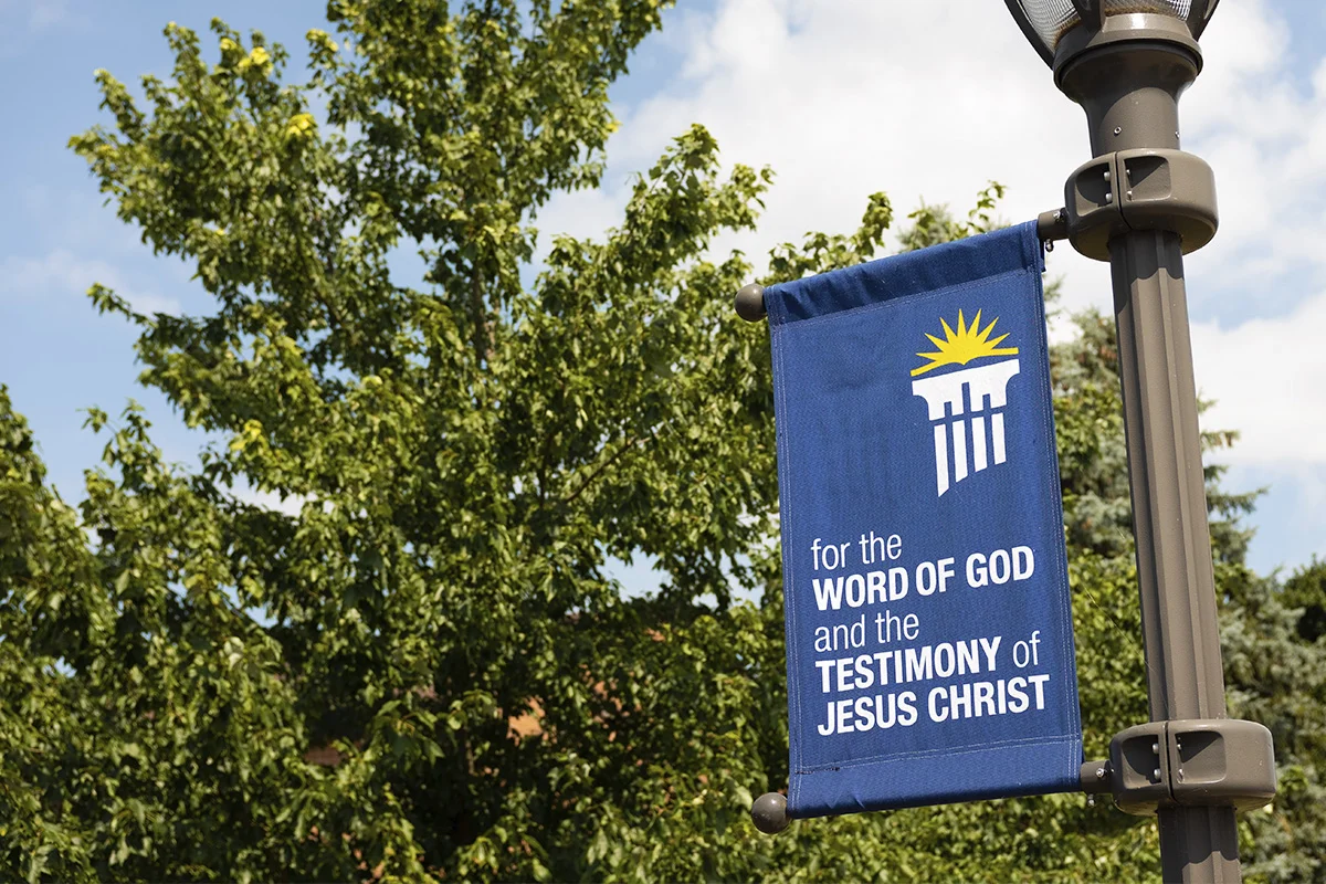 A vertical blue banner hanging from a street lamp pole featuring a yellow sunburst and white columns with a white outline of a book, with text that reads "for the WORD OF GOD and the TESTIMONY of JESUS CHRIST," against a background of green trees and