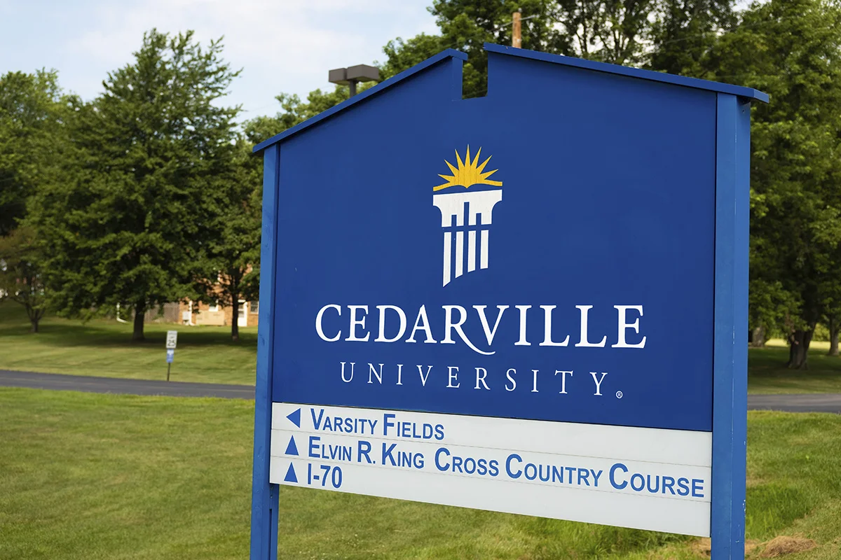 Signboard for Cedarville University with directions to Varsity Fields, Elvin R. King Cross Country Course, and I-70. Background shows green trees and grass.
