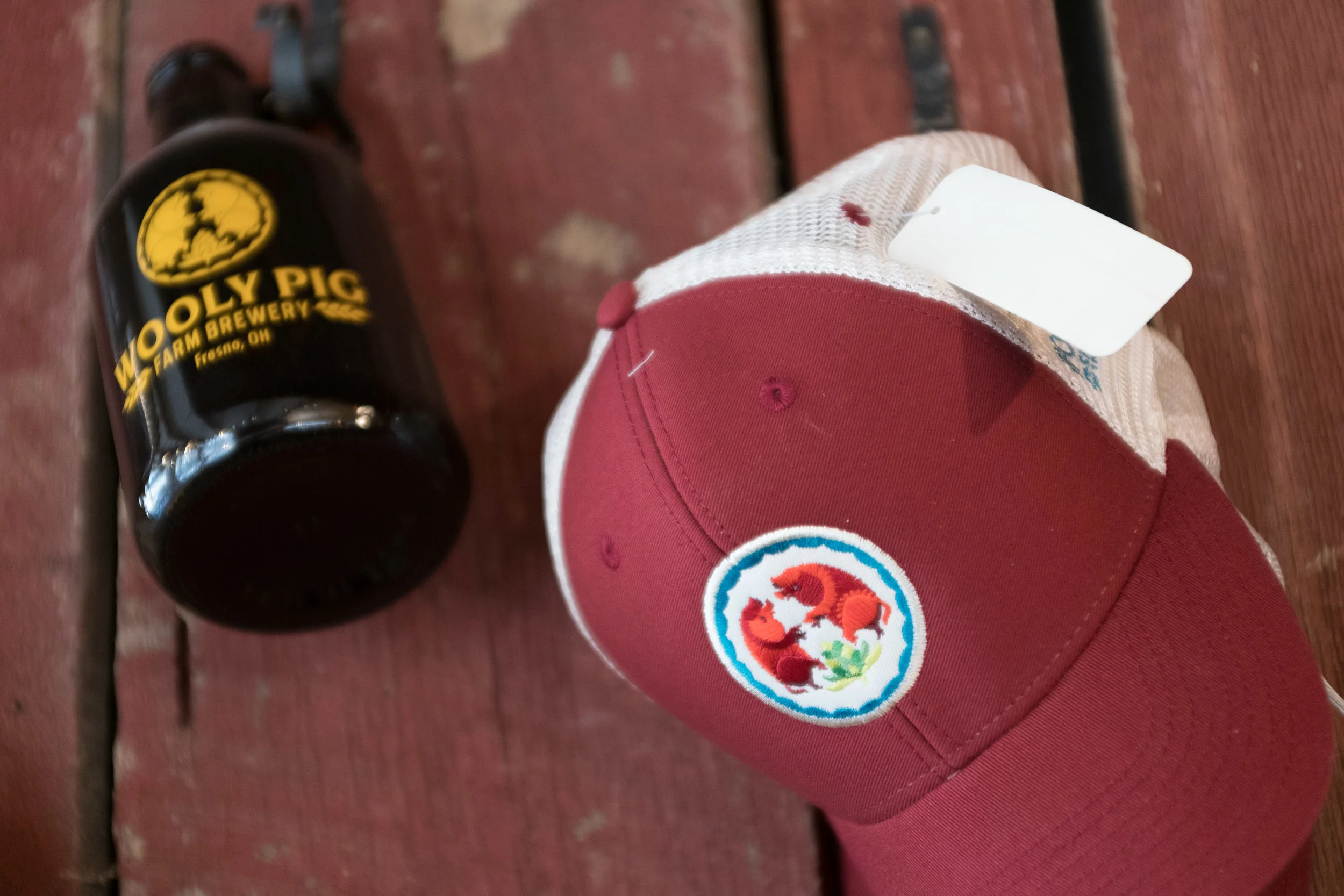 A red baseball cap with a colorful embroidered patch featuring two foxes, placed on a wooden surface next to a black mug with yellow logo for Wooly Pig Farm Brewery in Fresno, Ohio.