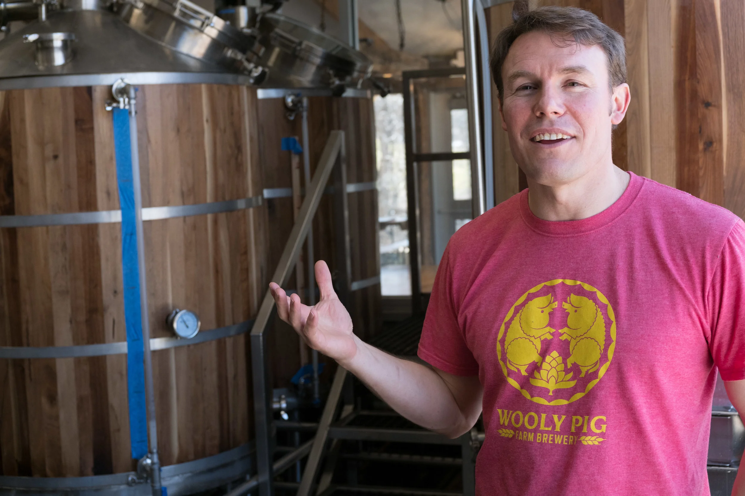 A man standing inside a brewery, gesturing with his hand towards brewing equipment, wearing a pink T-shirt with a yellow logo that says "Wooly Pig Farm Brewery."