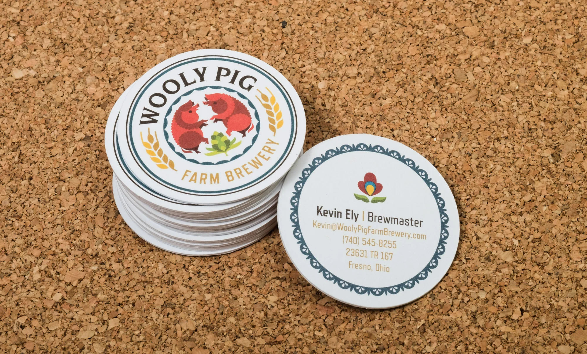 Business cards for Wooly Pig Farm Brewery featuring a logo with two red pigs and a business card with contact information for Kevin Ely, Brewmaster, on a cork surface.
